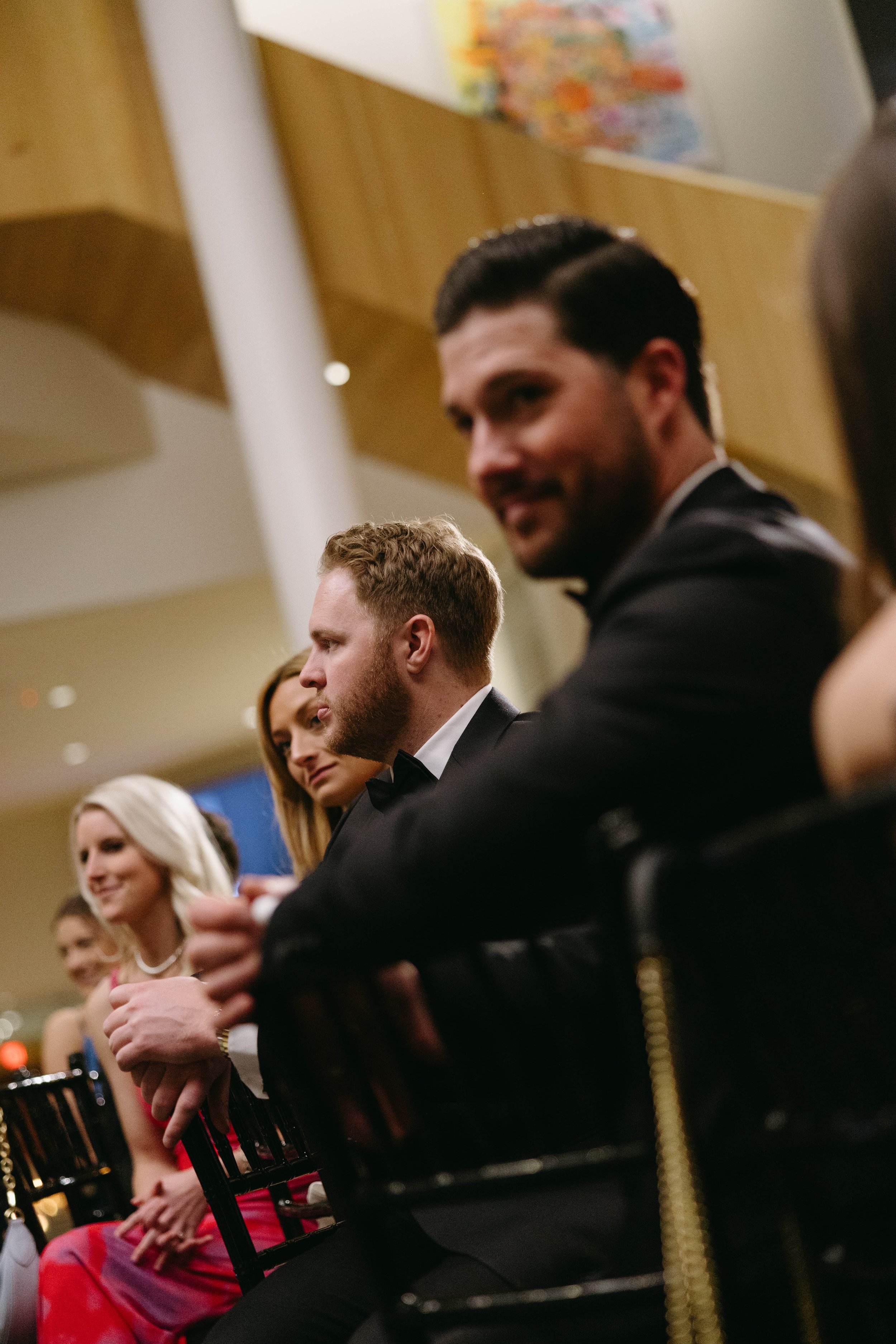 Group of well-dressed people sitting at an indoor event, with focus on a man in a tuxedo.
