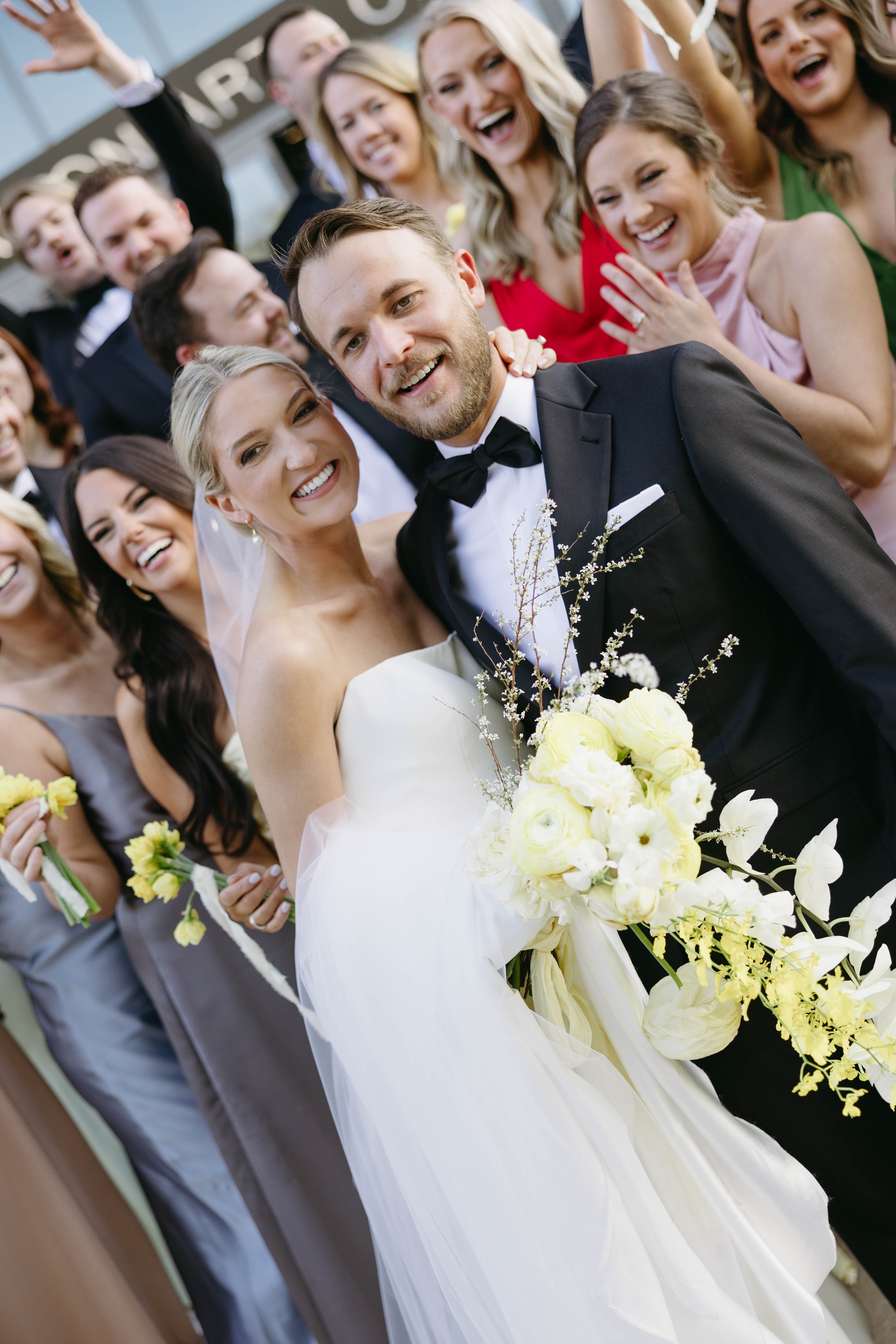 A joyful wedding party celebrating with a bride and groom. The bride wears a white dress and veil, holding a bouquet of yellow and white flowers. The groom is in a black tuxedo. Their friends surround them, smiling and cheering.