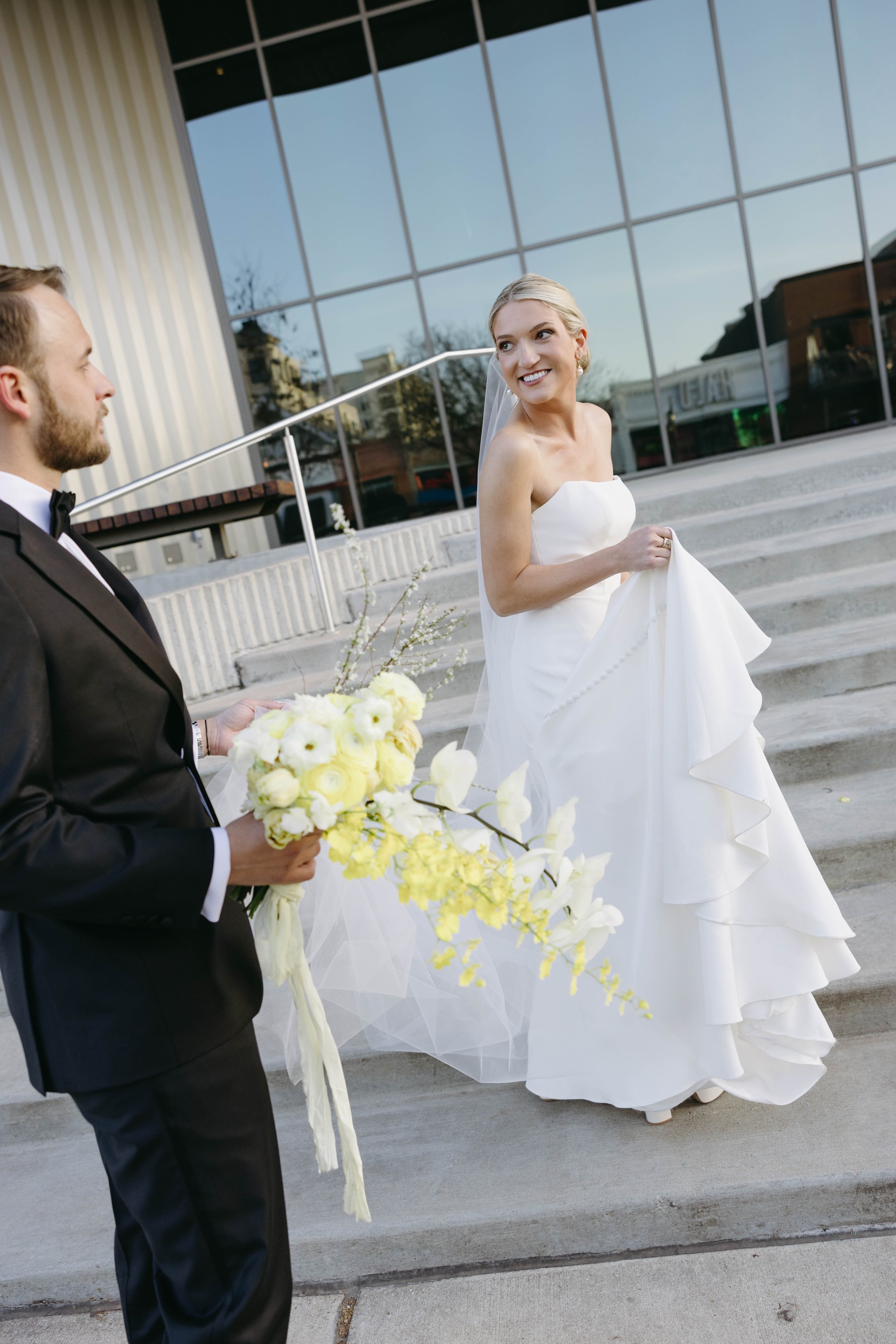 Bride and groom standing outside a building, bride in a white gown holding her dress, groom in a black suit holding a bouquet of yellow and white flowers; modern glass building in background.