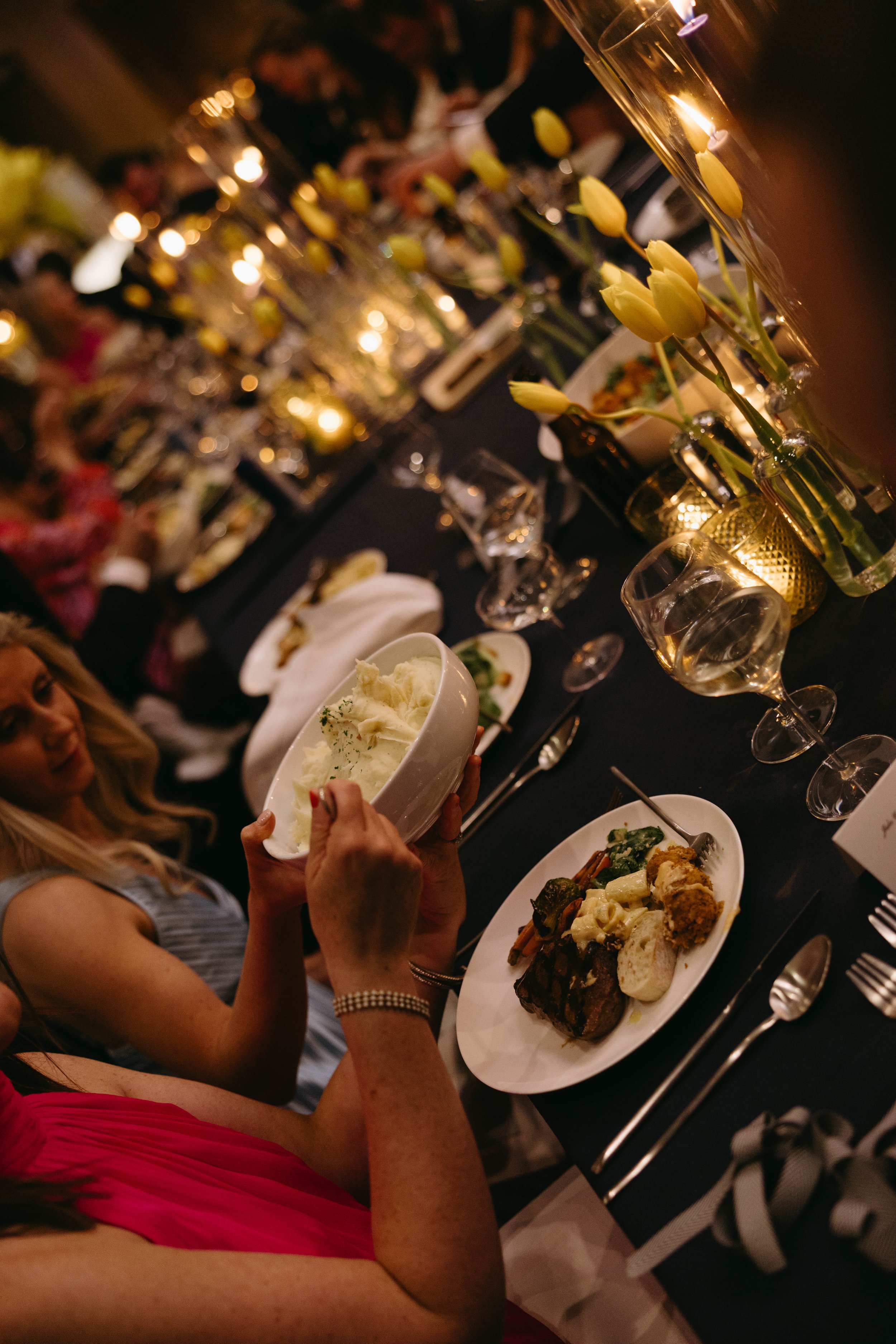 Elegant dinner table setting with guests, featuring plates of food, yellow tulip decorations, and candlelight.