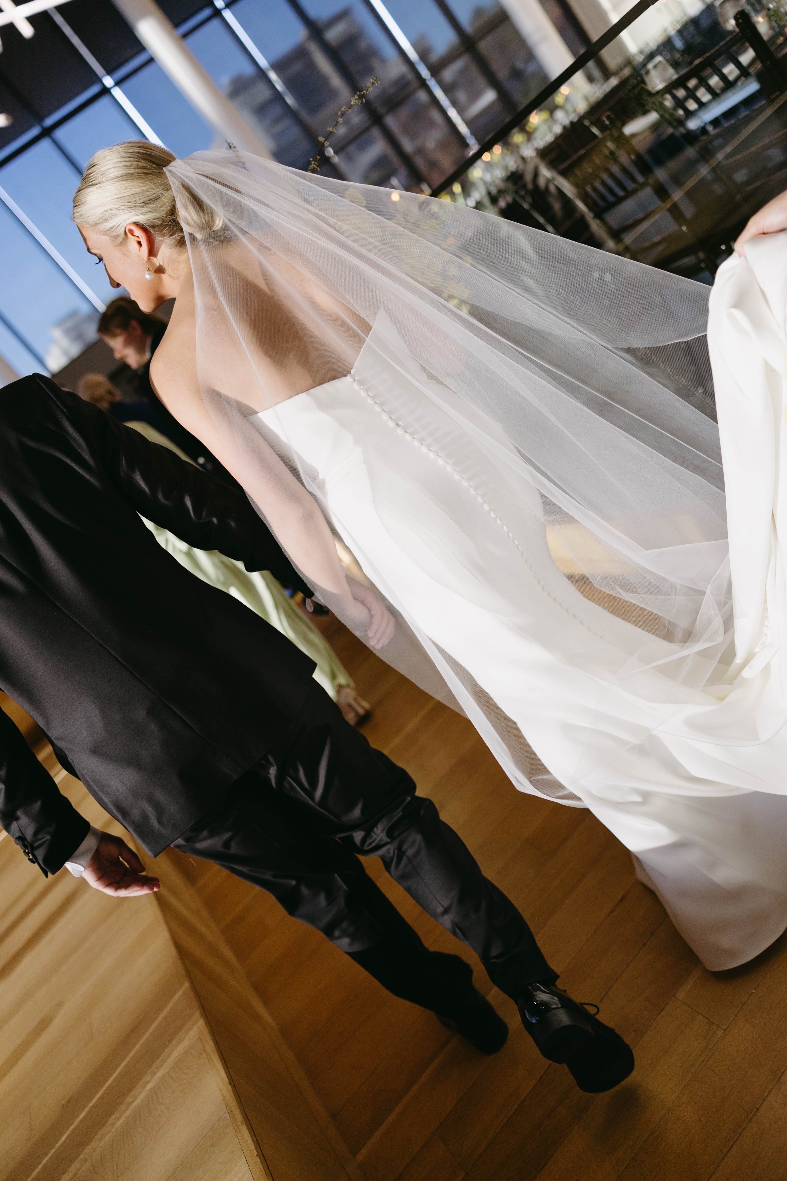 Bride in white strapless wedding gown with long veil, walking indoors alongside a person in a black suit.