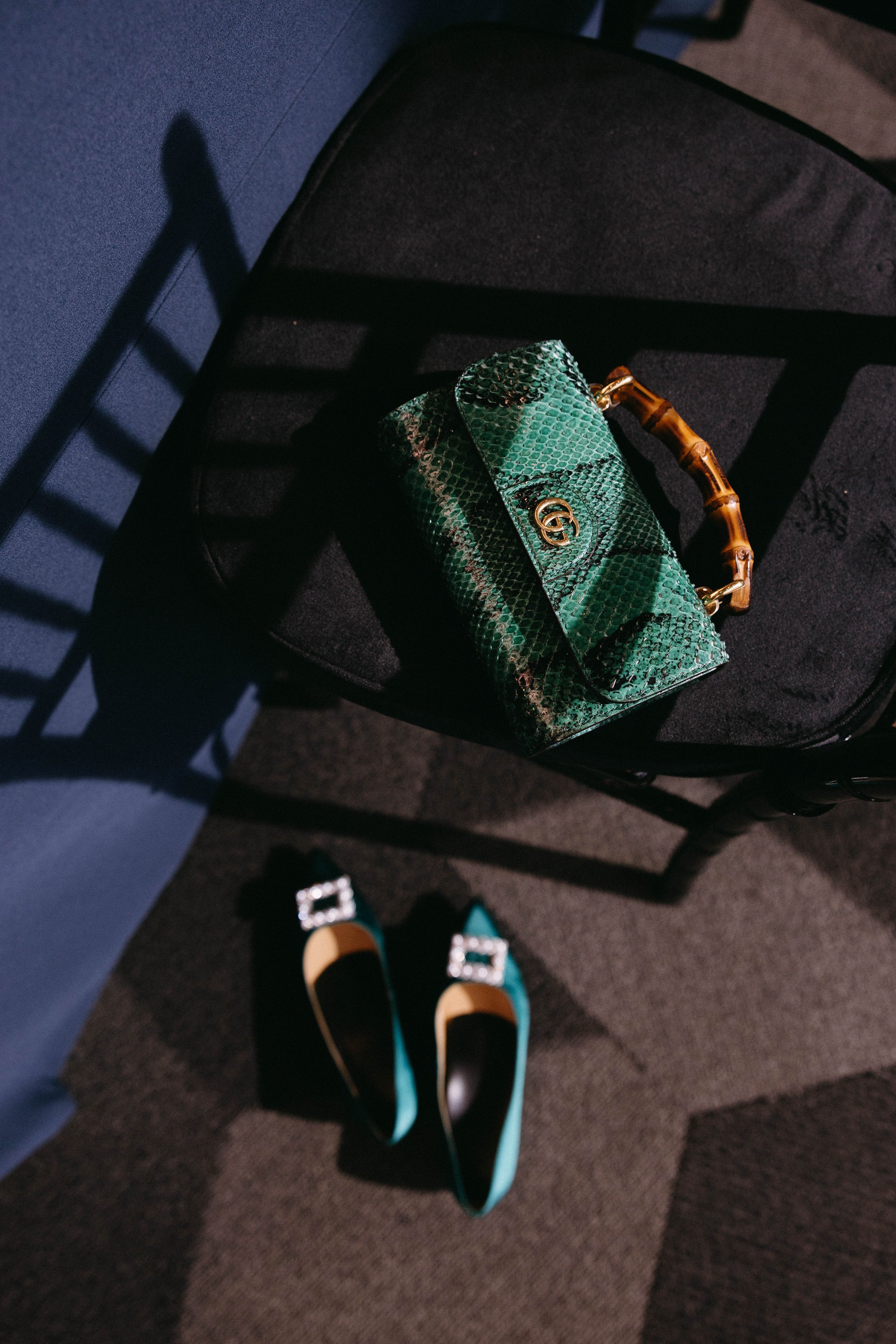 A green snakeskin handbag with a bamboo handle on a black chair, paired with teal high-heeled shoes with rhinestone buckles on a checkered carpet.