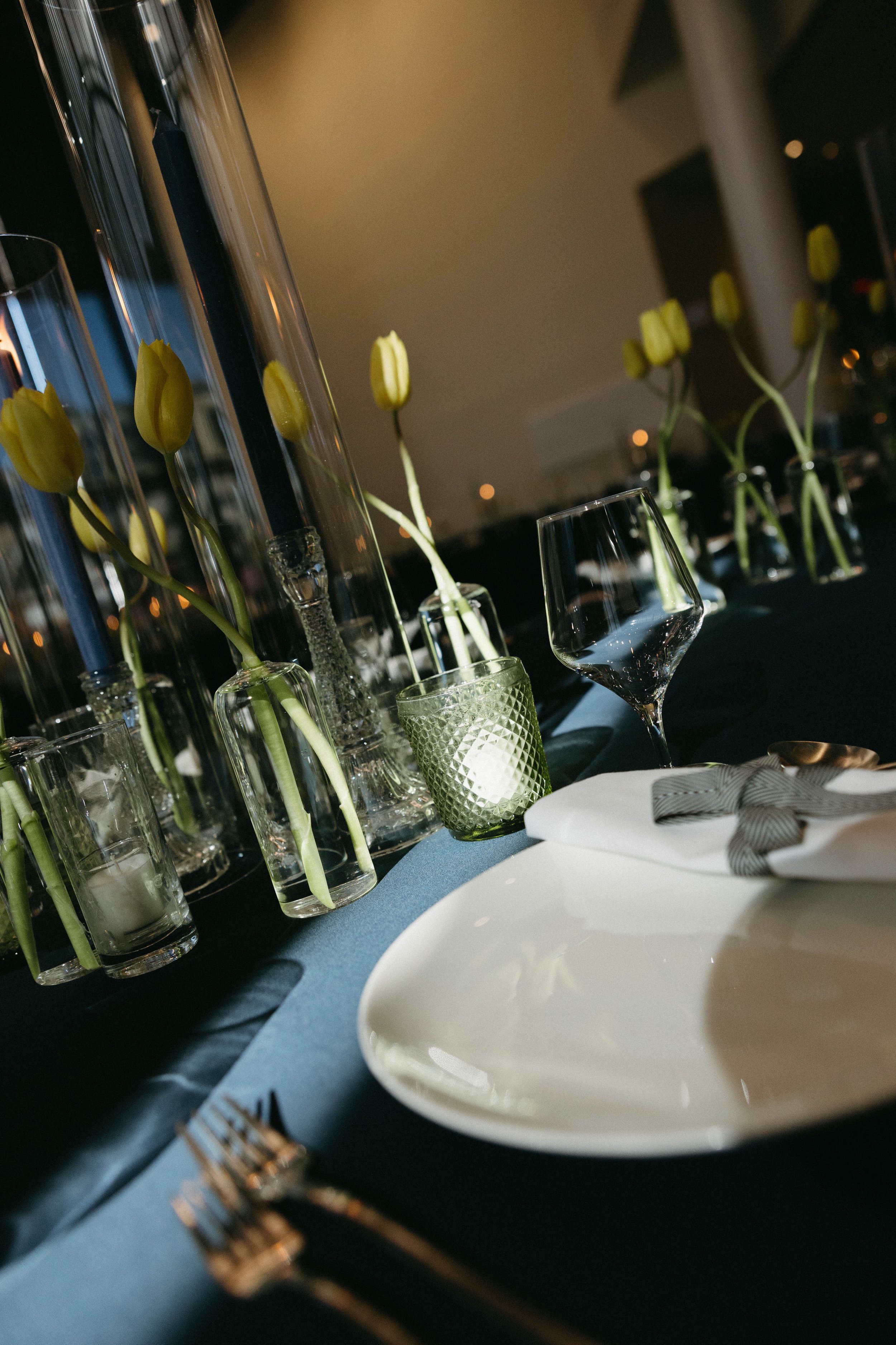 Elegant table setting with yellow tulips, glass candle holders, wine glass, and crockery on a dark tablecloth.