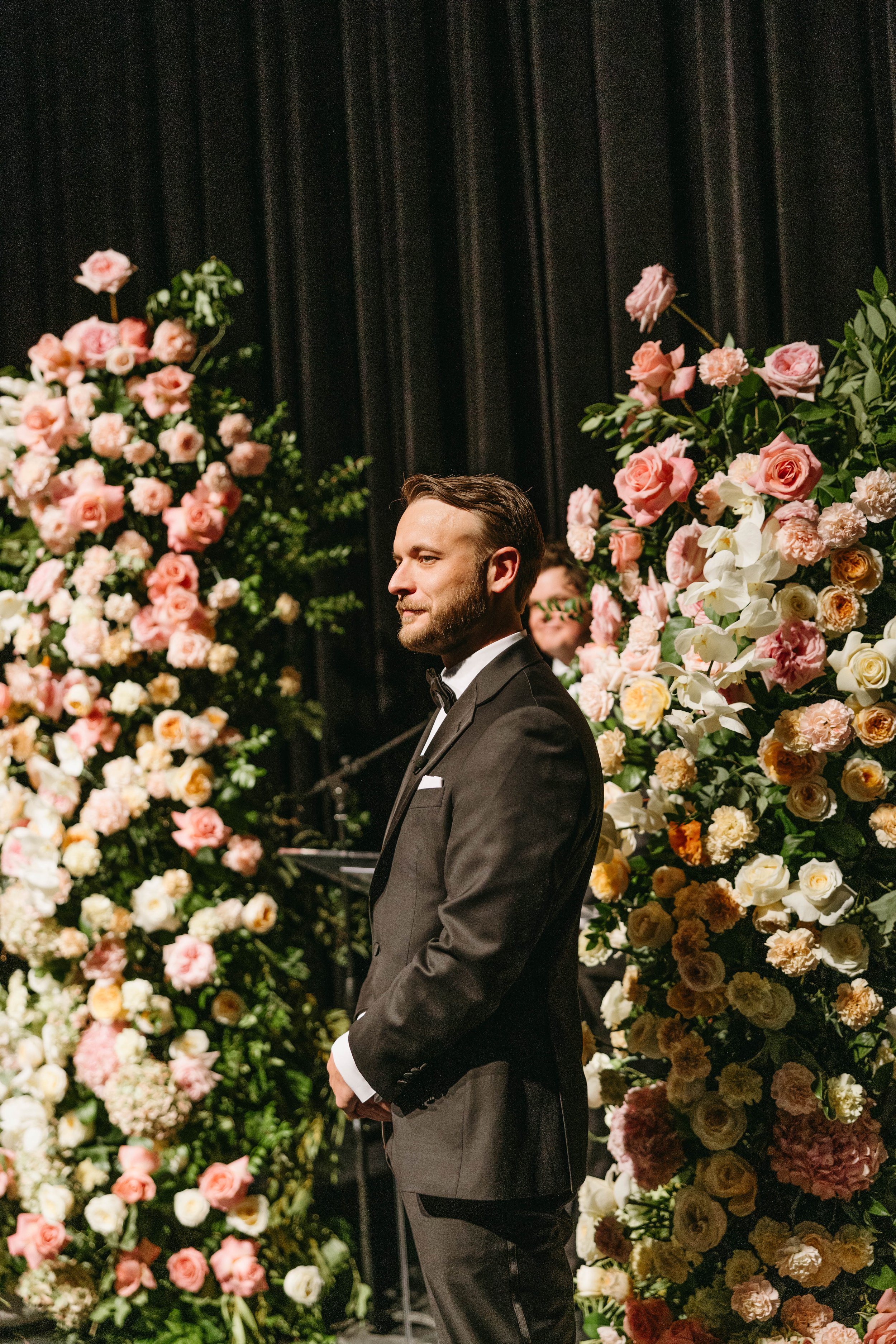 Man in a suit standing next to colorful flower arrangements at an indoor event.
