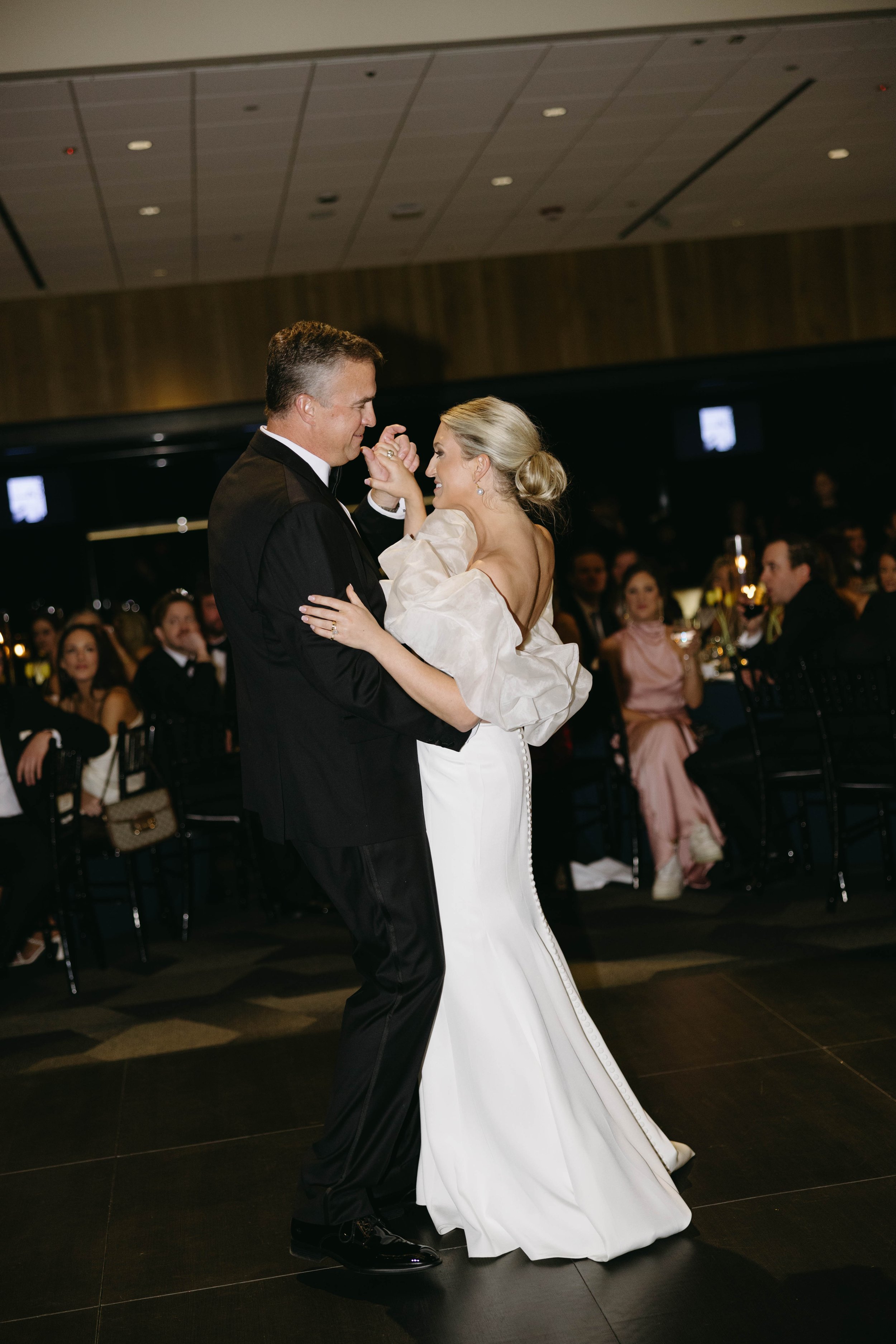 A couple dancing at a formal event with guests seated in the background.