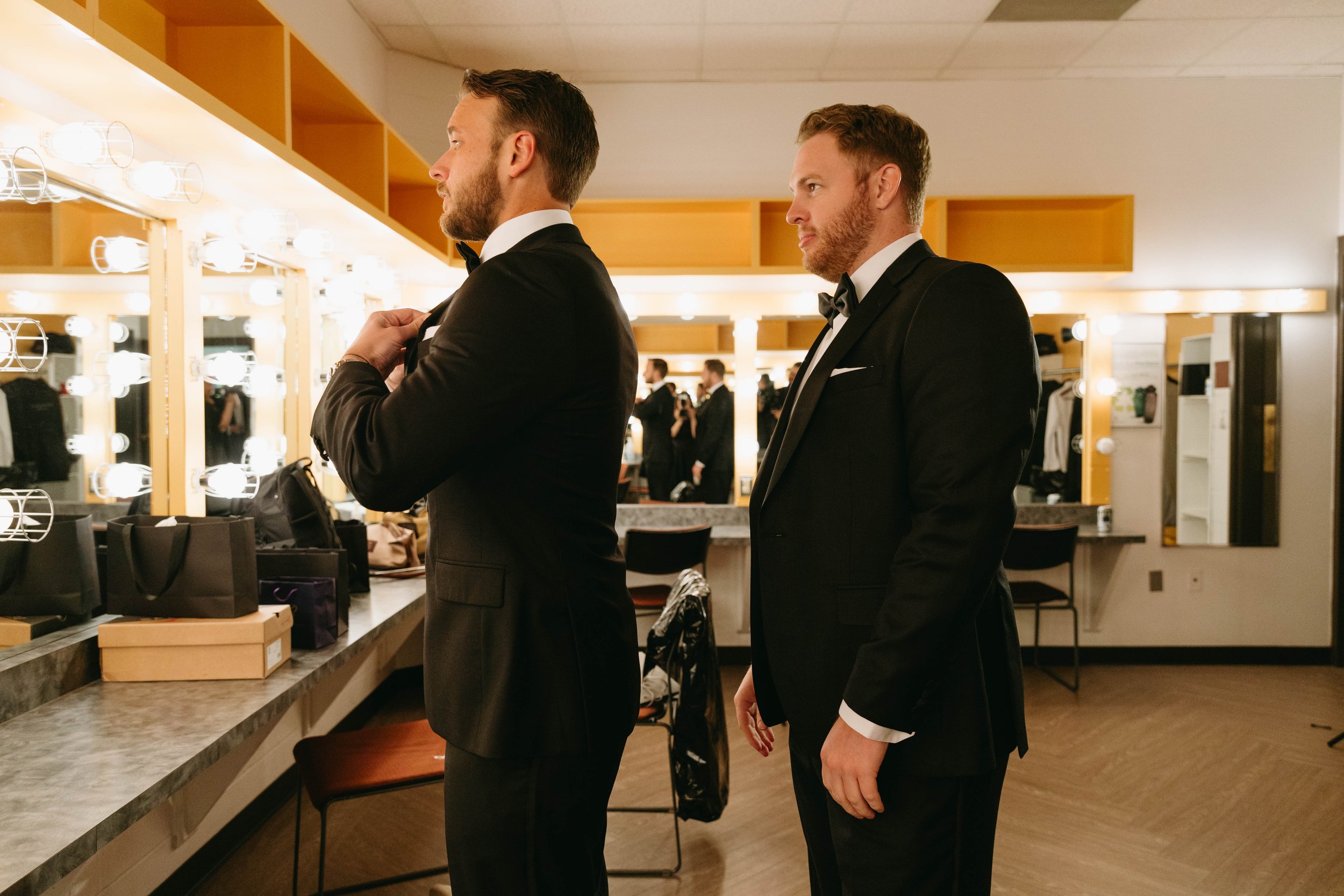 Two men in suits adjusting their bow ties in a dressing room with mirrors and bright vanity lights.