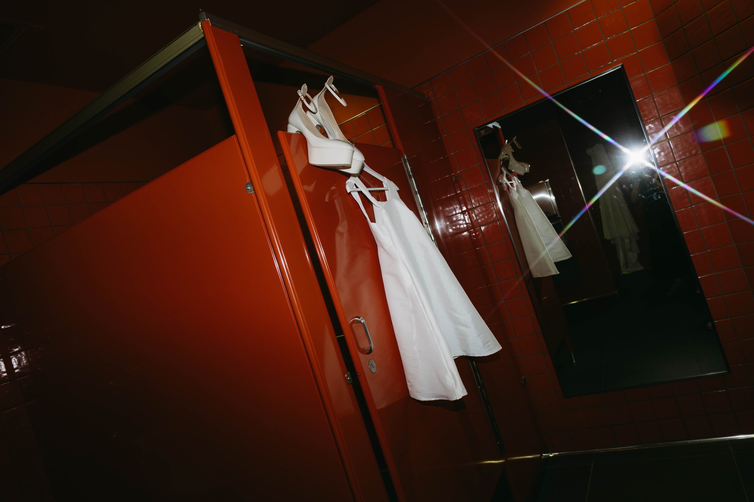 White dress and heels hanging on a red bathroom stall door, reflective mirror surface showing partial reflection.