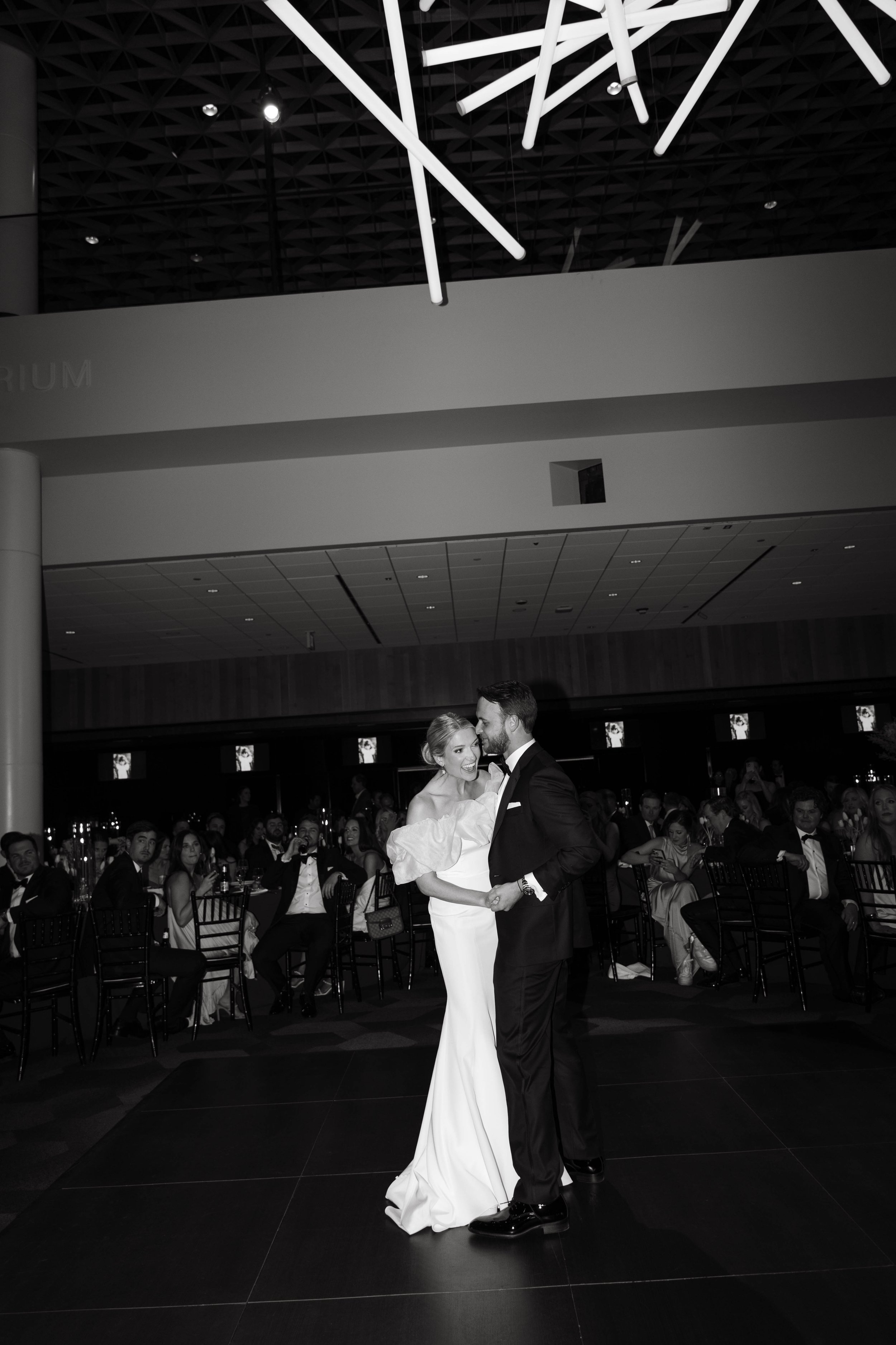 A black and white photo of a couple dancing at a formal event with guests seated in the background.