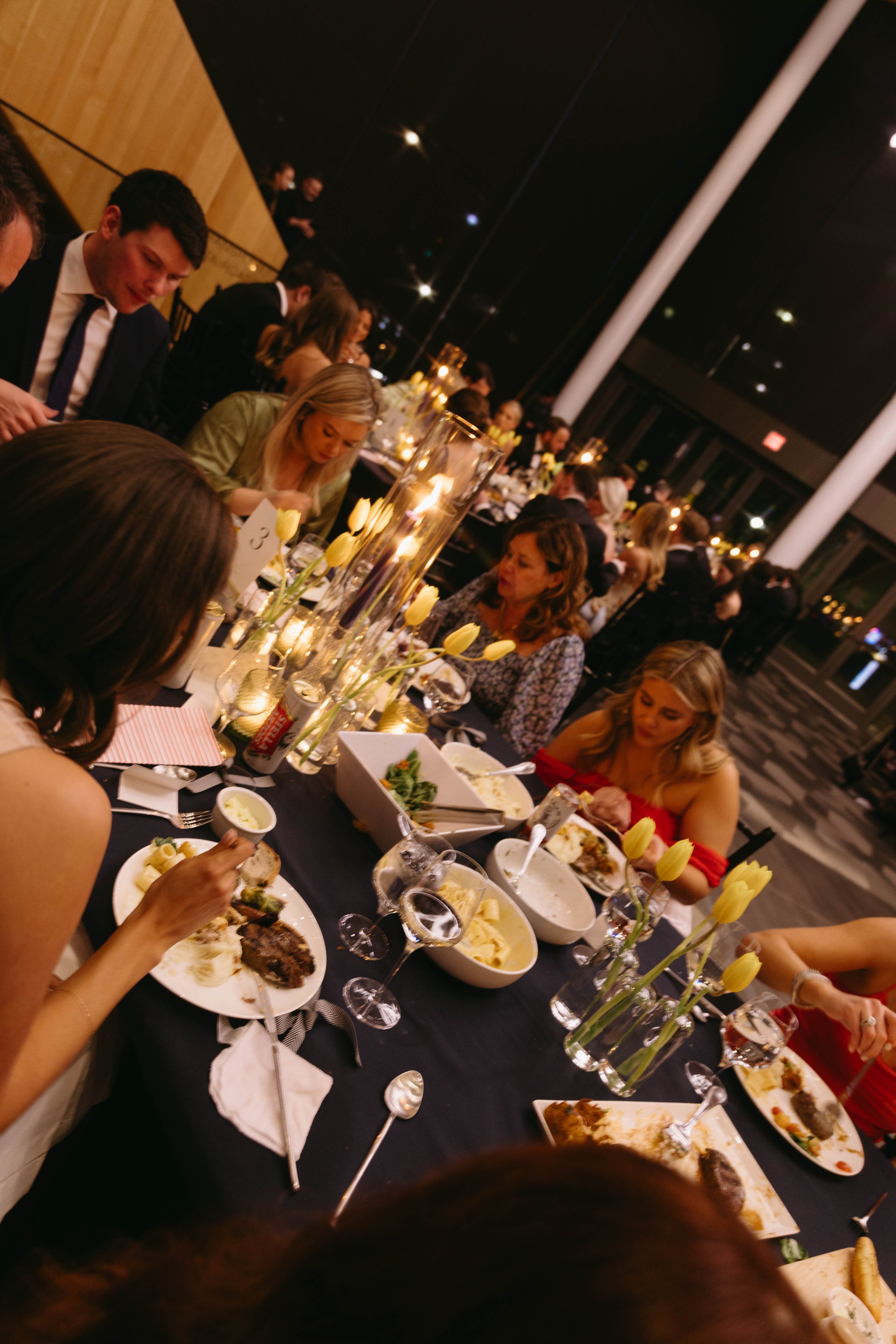 People seated at a formal dinner event, eating and conversing at a table set with flowers, candles, and various dishes in an elegant venue.