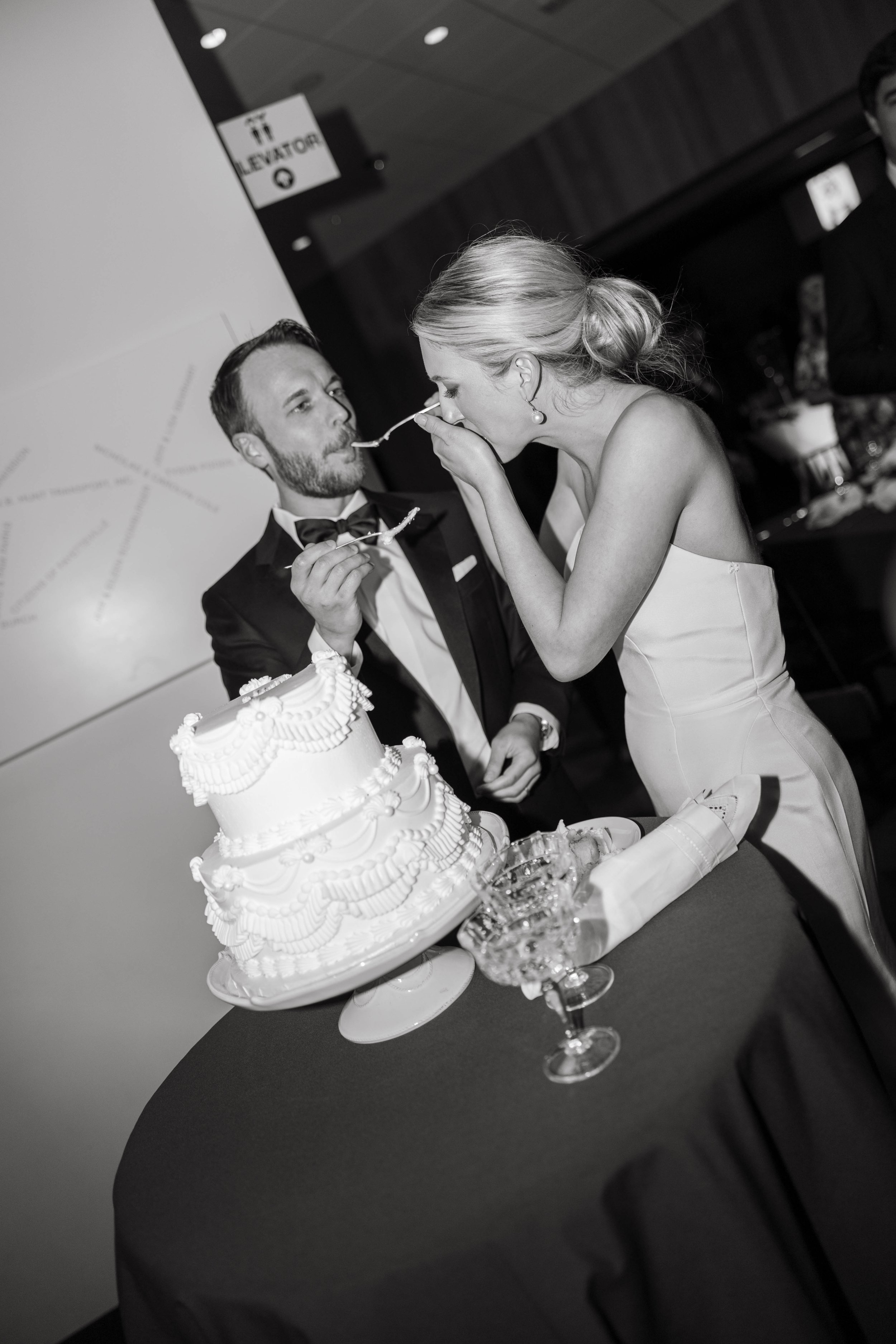 A bride and groom feeding each other cake at their wedding, standing next to a tiered wedding cake on a table.