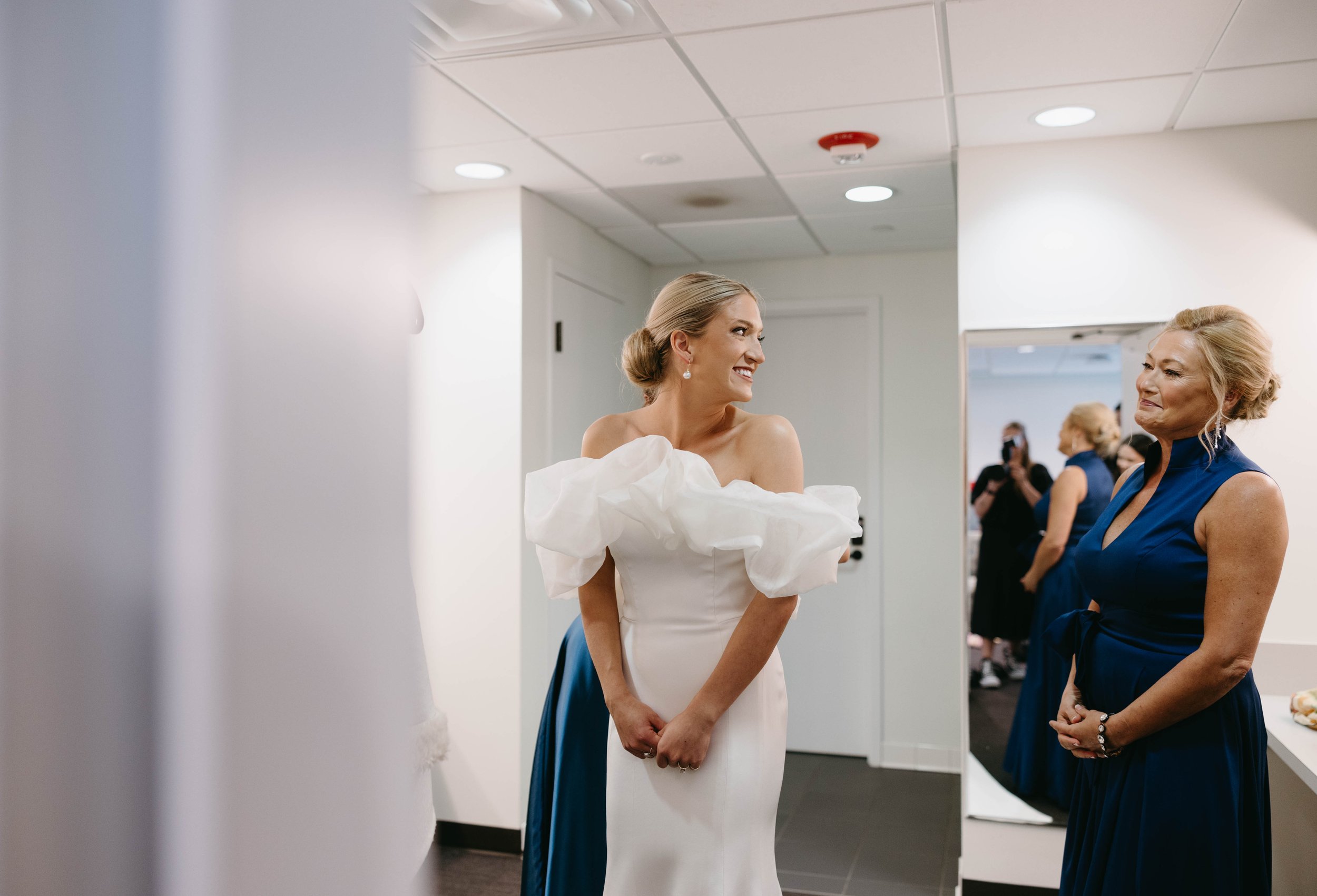 Bride in white dress with ruffled sleeves smiling, standing beside a woman in a blue dress in a dressing room.
