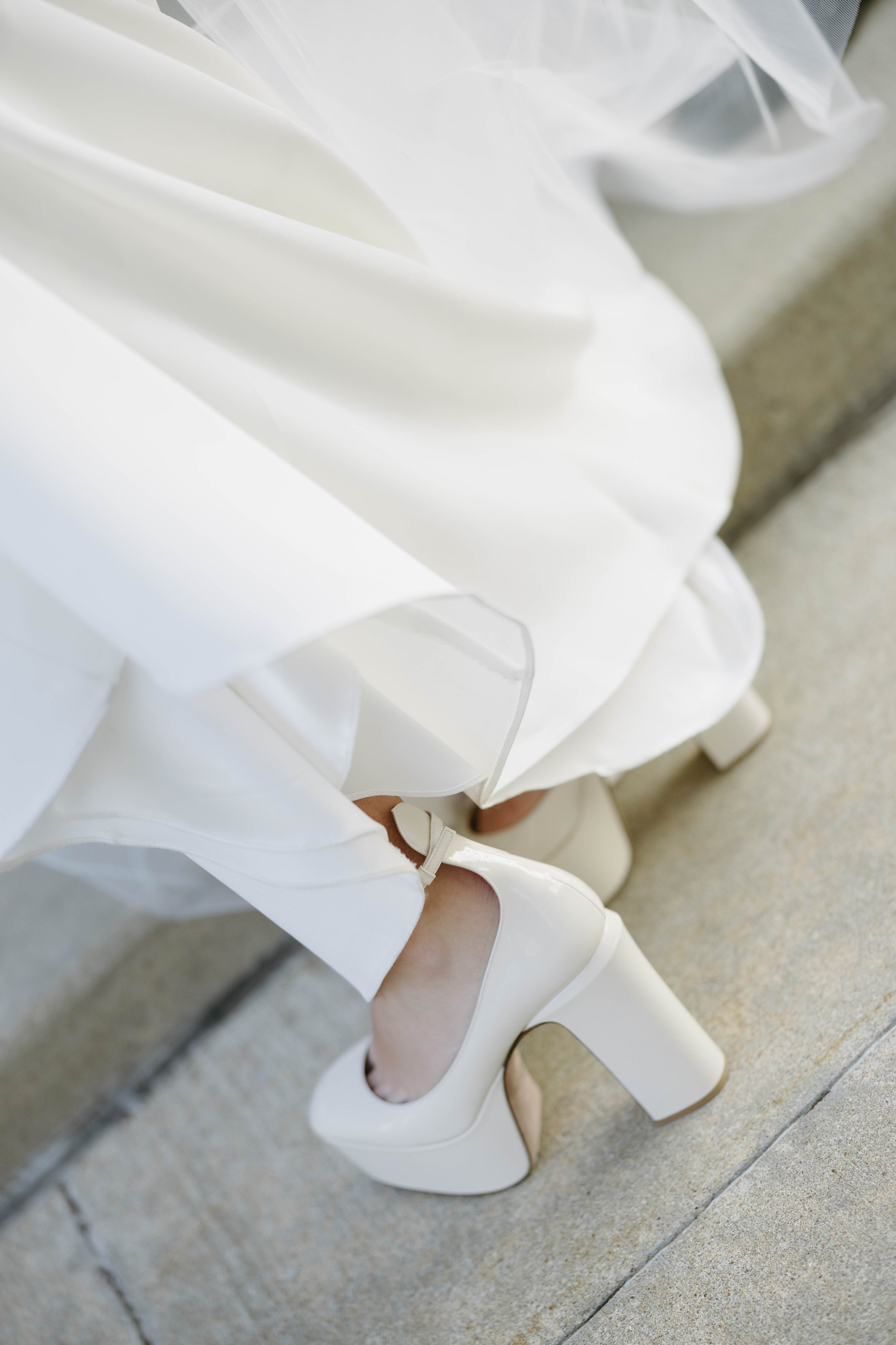 Close-up of a person wearing white platform high heels and a flowing white dress, standing on concrete steps.