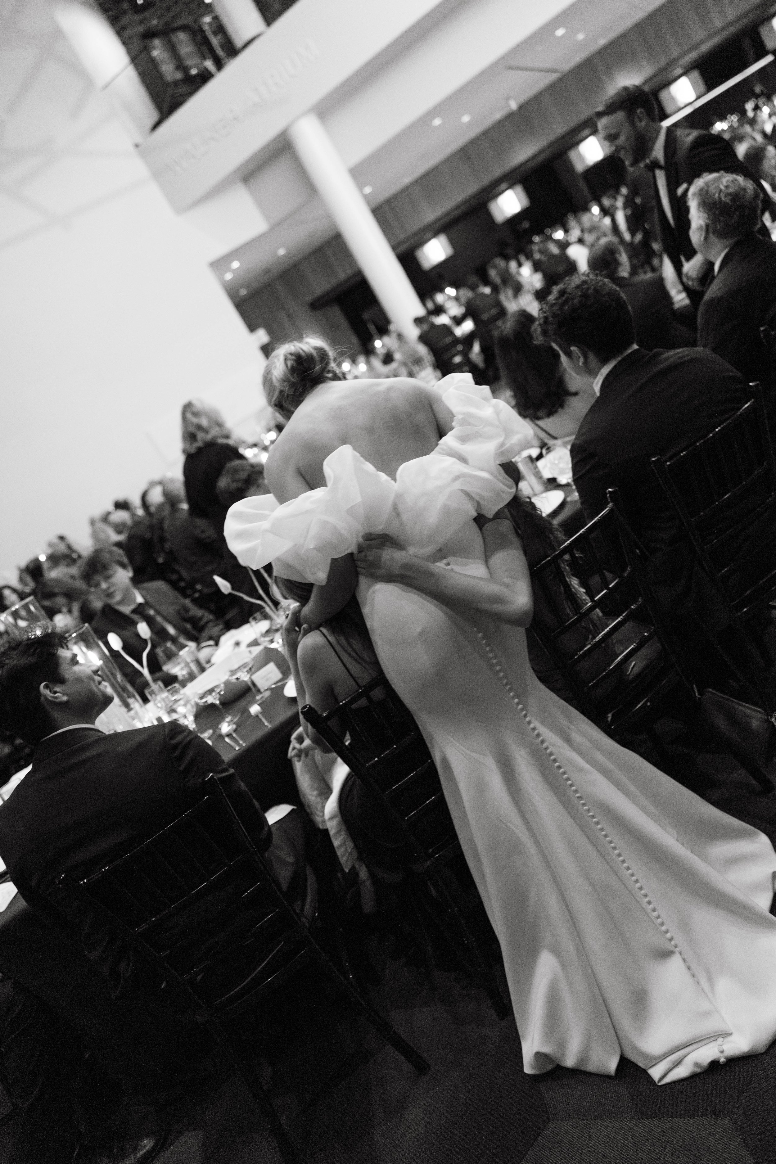 Elegant event with guests seated at round tables, woman in a gown hugging another seated woman, black and white photo.