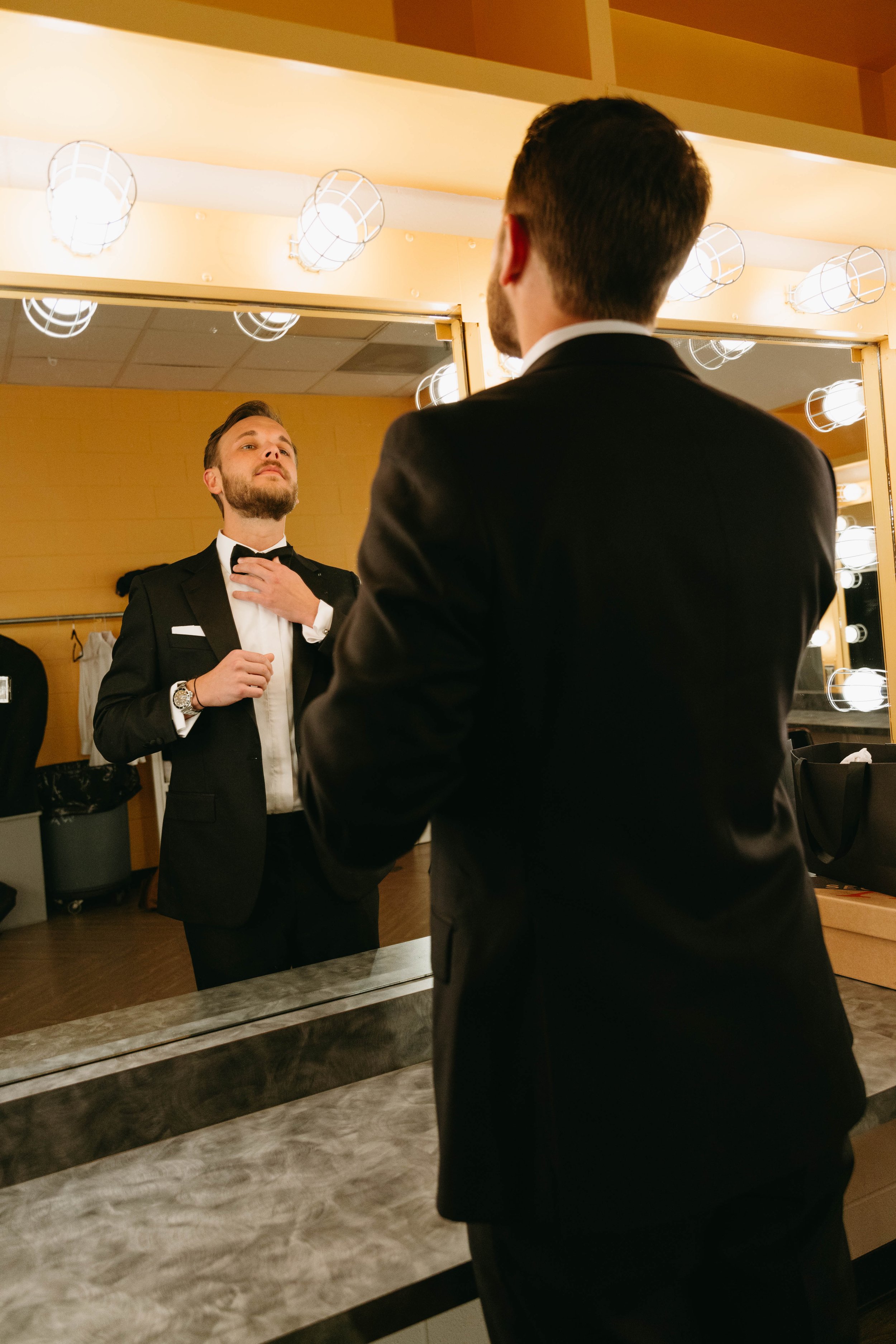 Man in a suit adjusting a bow tie in front of a mirror with bright lights in a dressing room.