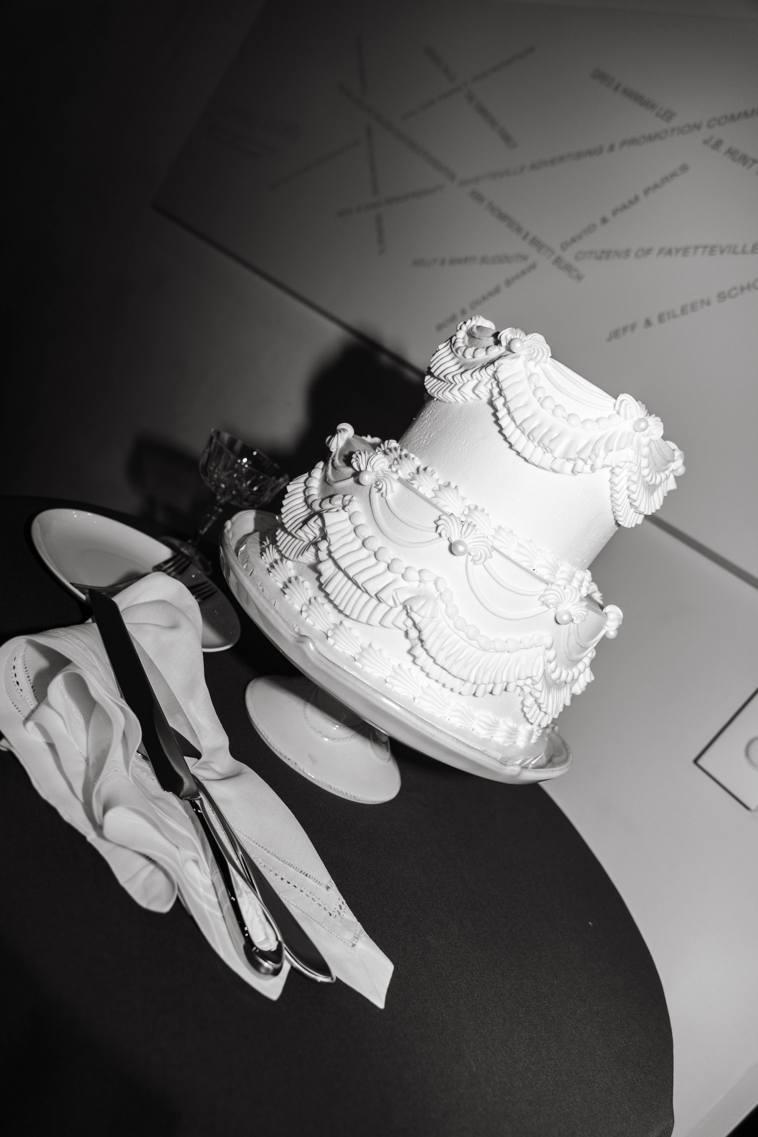 Black and white photo of a tiered white cake with intricate decorative icing placed on a cake stand next to a folded napkin and cutlery.