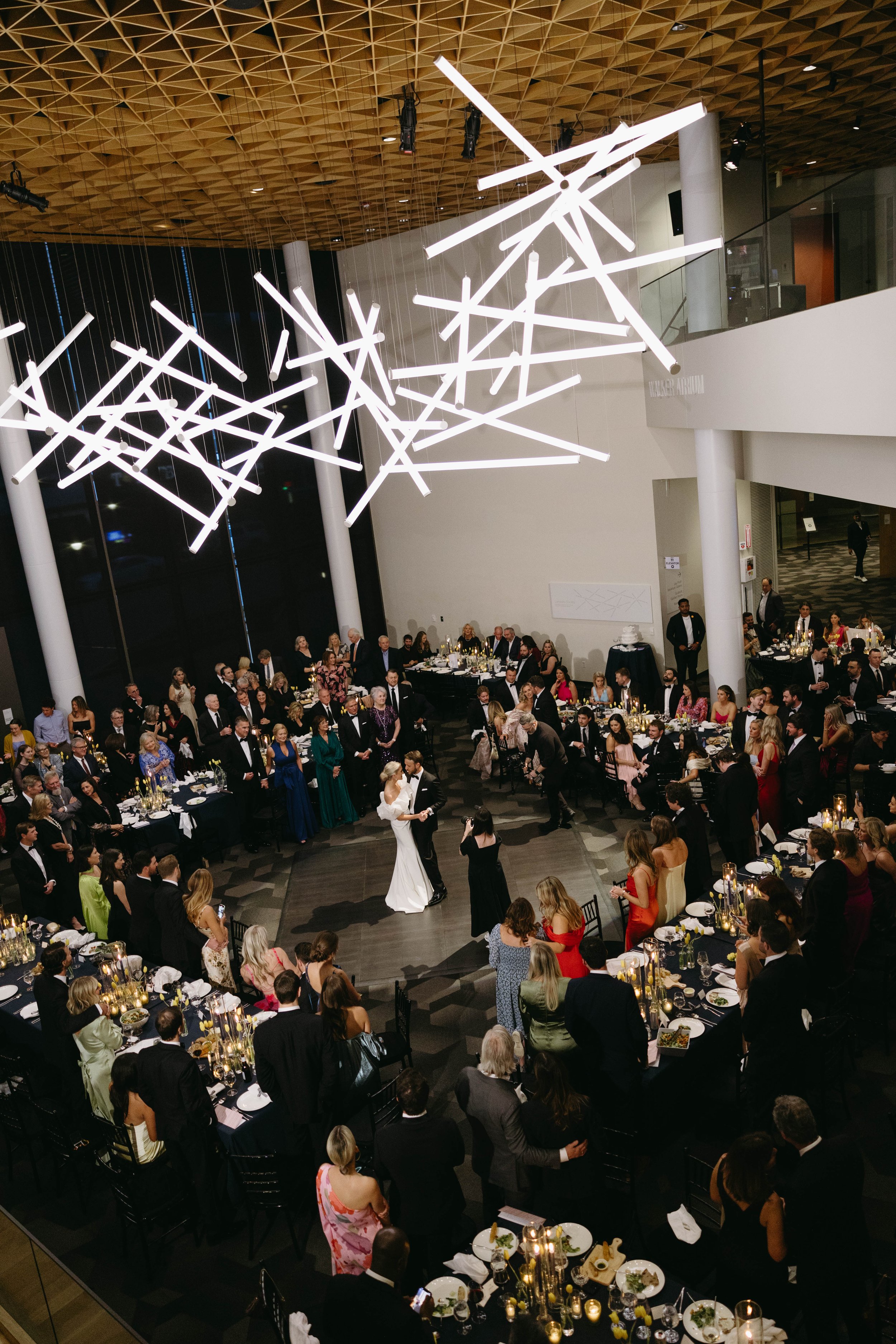 Aerial view of a wedding reception with guests seated at tables and a couple dancing under modern chandeliers.