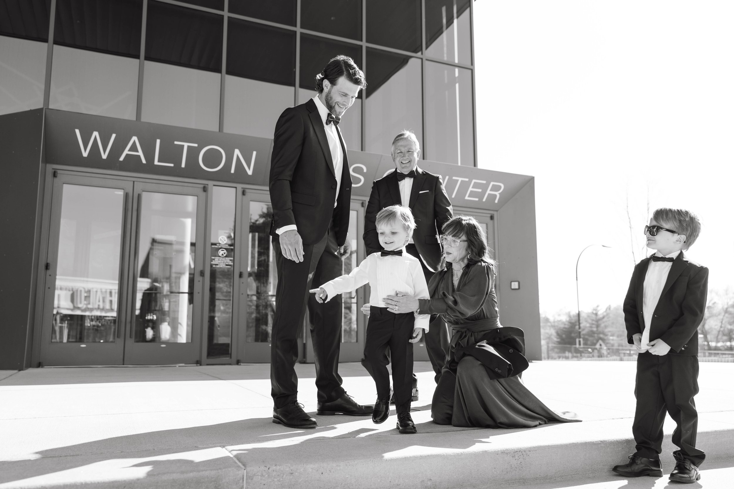 A group of people dressed in formal attire stands outside the Walton Arts Center. An adult and a child are prominent in formal suits, with a woman kneeling to assist the child. Another child in a suit and sunglasses stands nearby. The mood appears jo