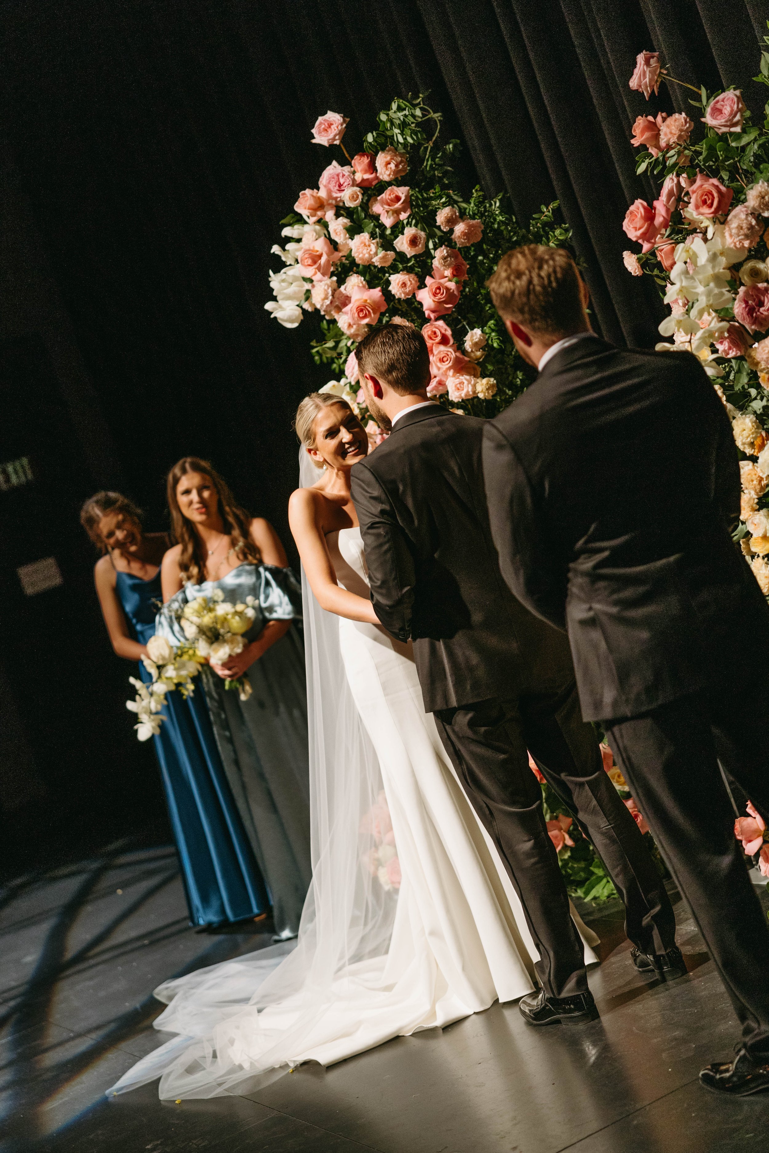 A bride and groom exchanging vows during a wedding ceremony, surrounded by floral arrangements, with bridesmaids in the background holding bouquets.