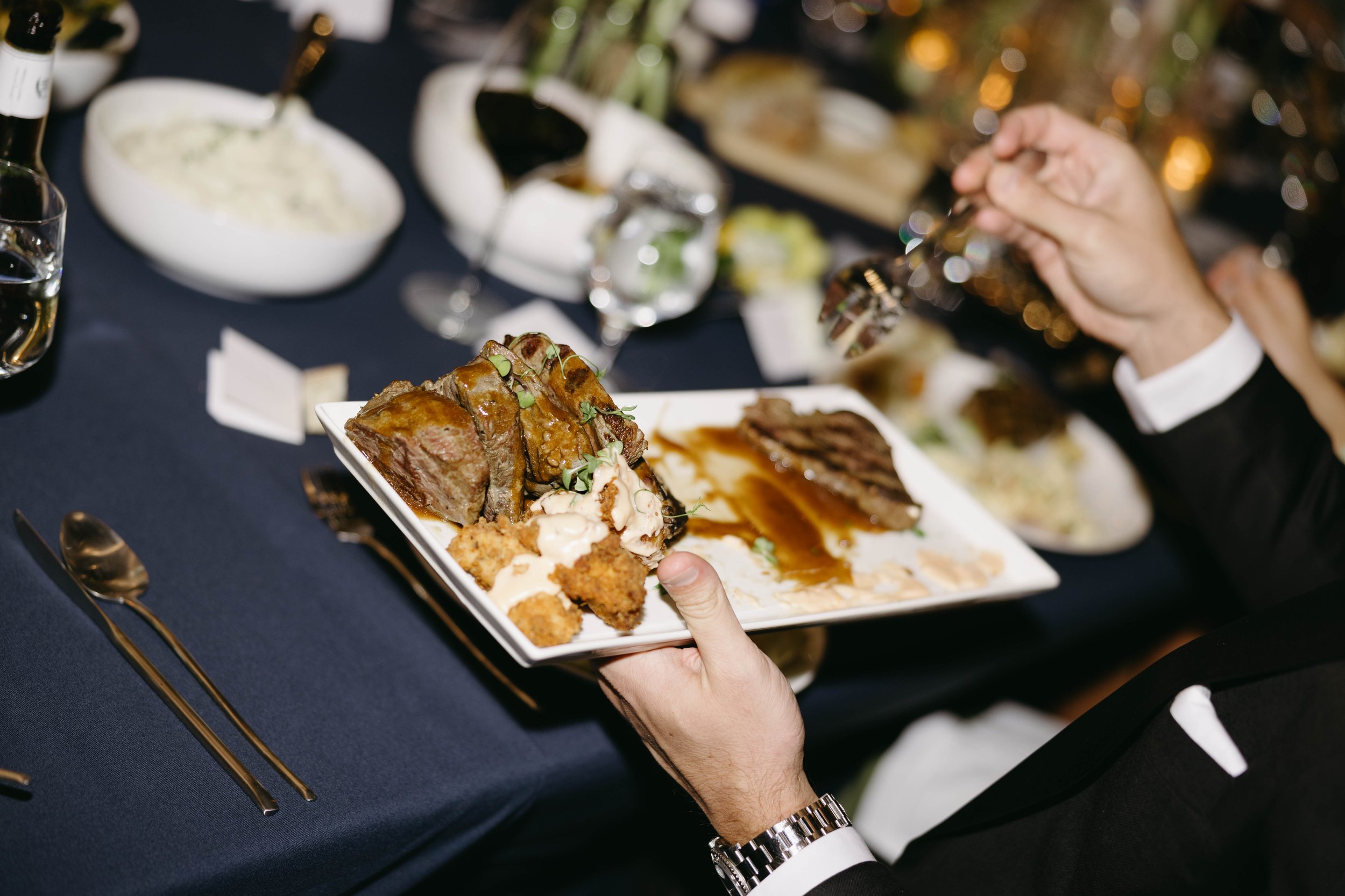 Person holding a plate of gourmet steak, garnished with herbs, sauce, and side dishes at a formal dinner setting with blurred background.