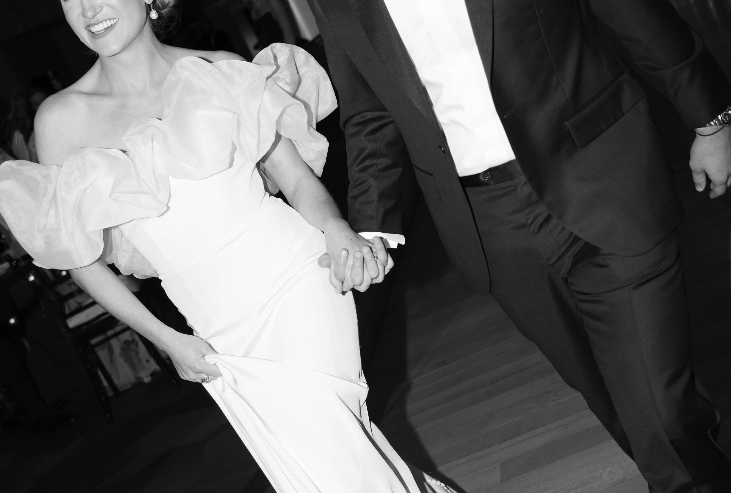 A couple in formal attire holding hands, the woman in a white dress with ruffled sleeves, and the man in a suit, smiling and walking indoors.