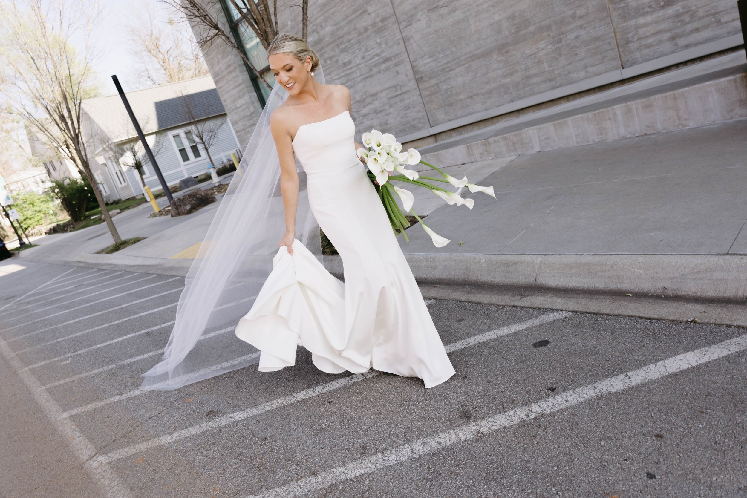 Bride in white wedding dress holding calla lilies, walking in a parking lot, with veil flowing.