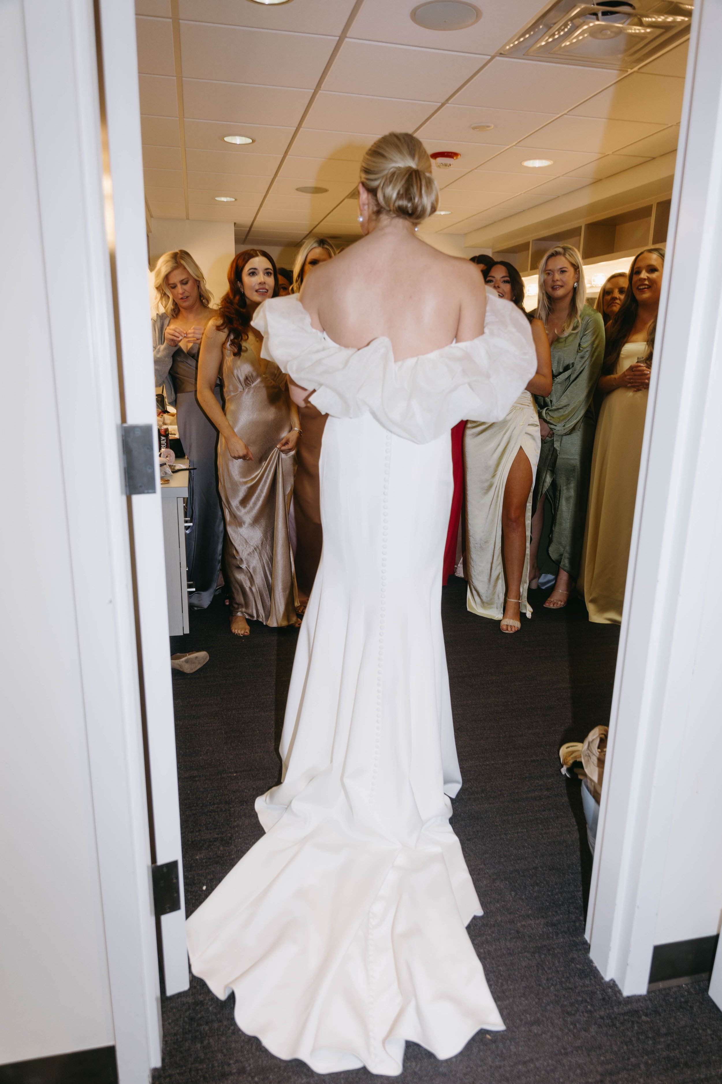 A bride in an off-shoulder wedding dress stands with bridesmaids in different colored dresses in a room.