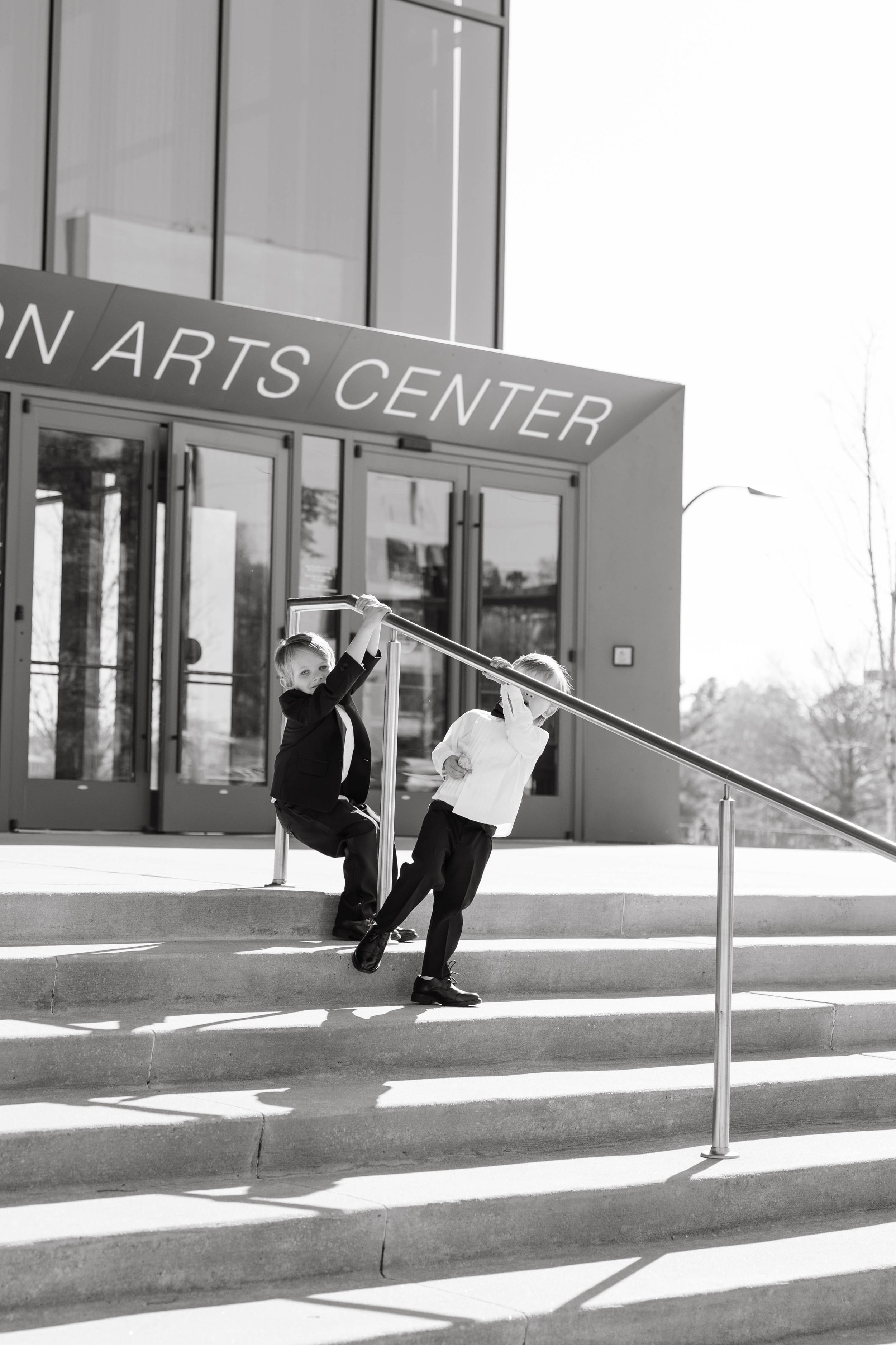 Two children playing on a staircase handrail in front of a building with the sign 'ARTS CENTER'. The kids are dressed in formal attire.