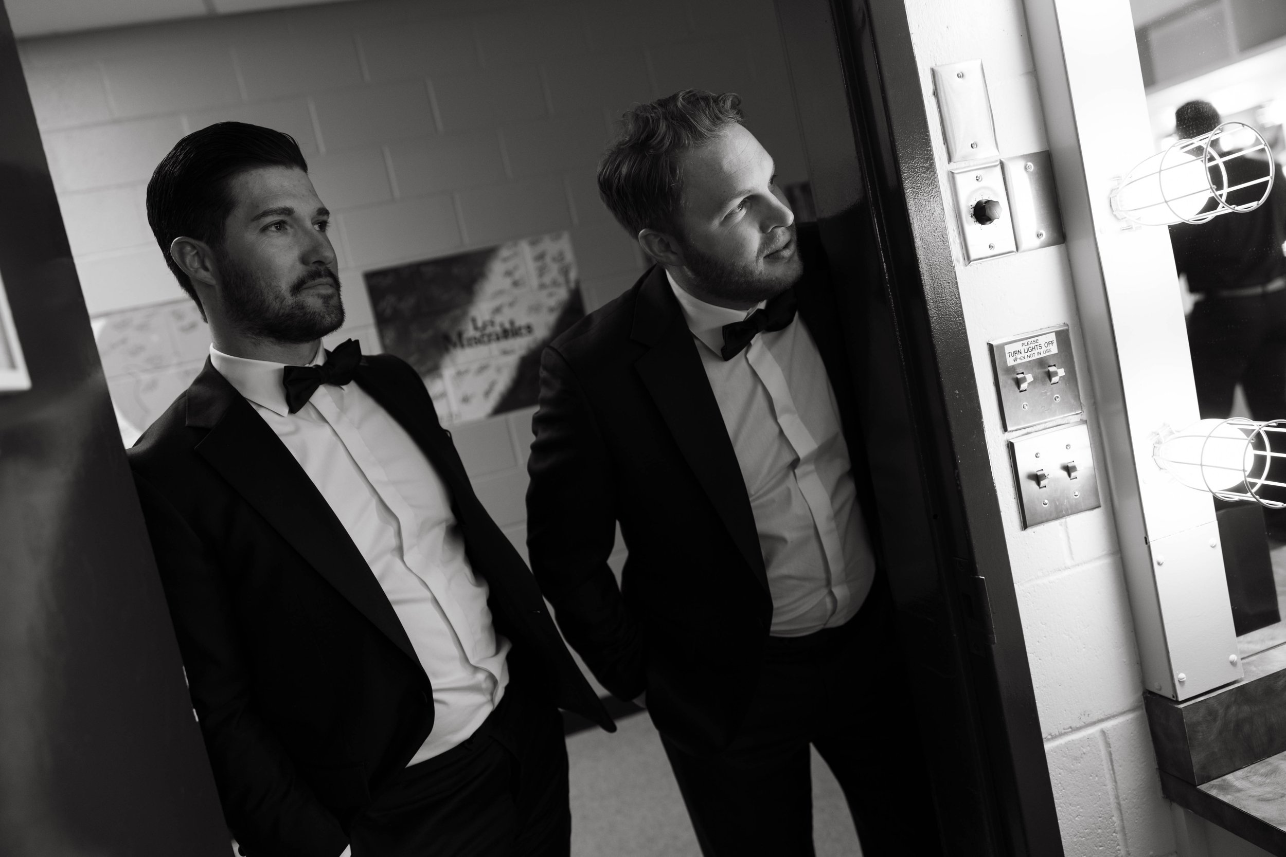 Two men in formal attire, wearing tuxedos with bow ties, stand near a doorway, one looking at a dressing room mirror lined with lights. A "Les Miserables" poster is partially visible on the wall in the background.