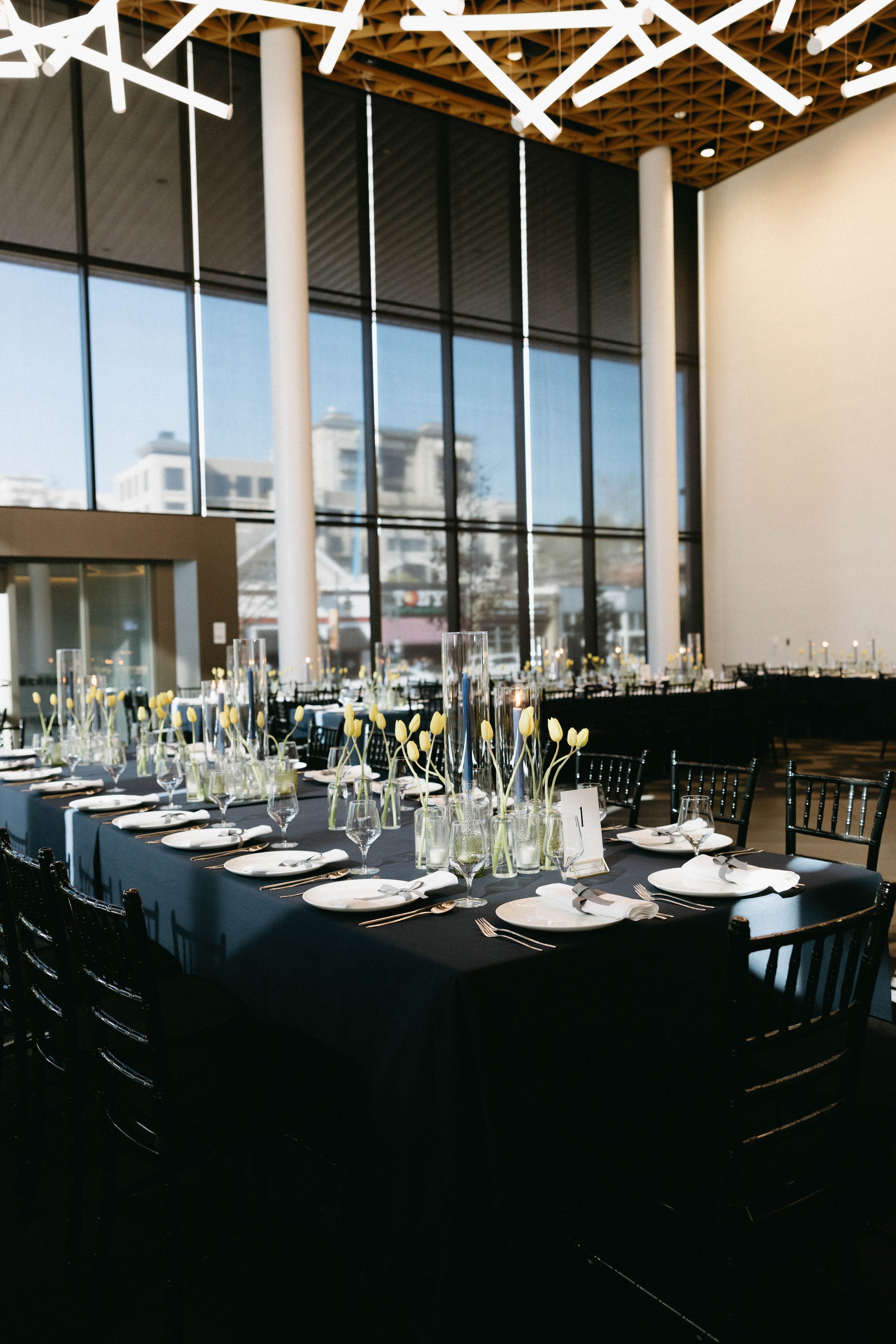 Elegantly set dining tables in a large, modern room with high ceilings and large windows. The tables are covered with black tablecloths, arranged with white plates, glasses, and cutlery. Yellow tulips in tall vases serve as centerpieces. Black chairs