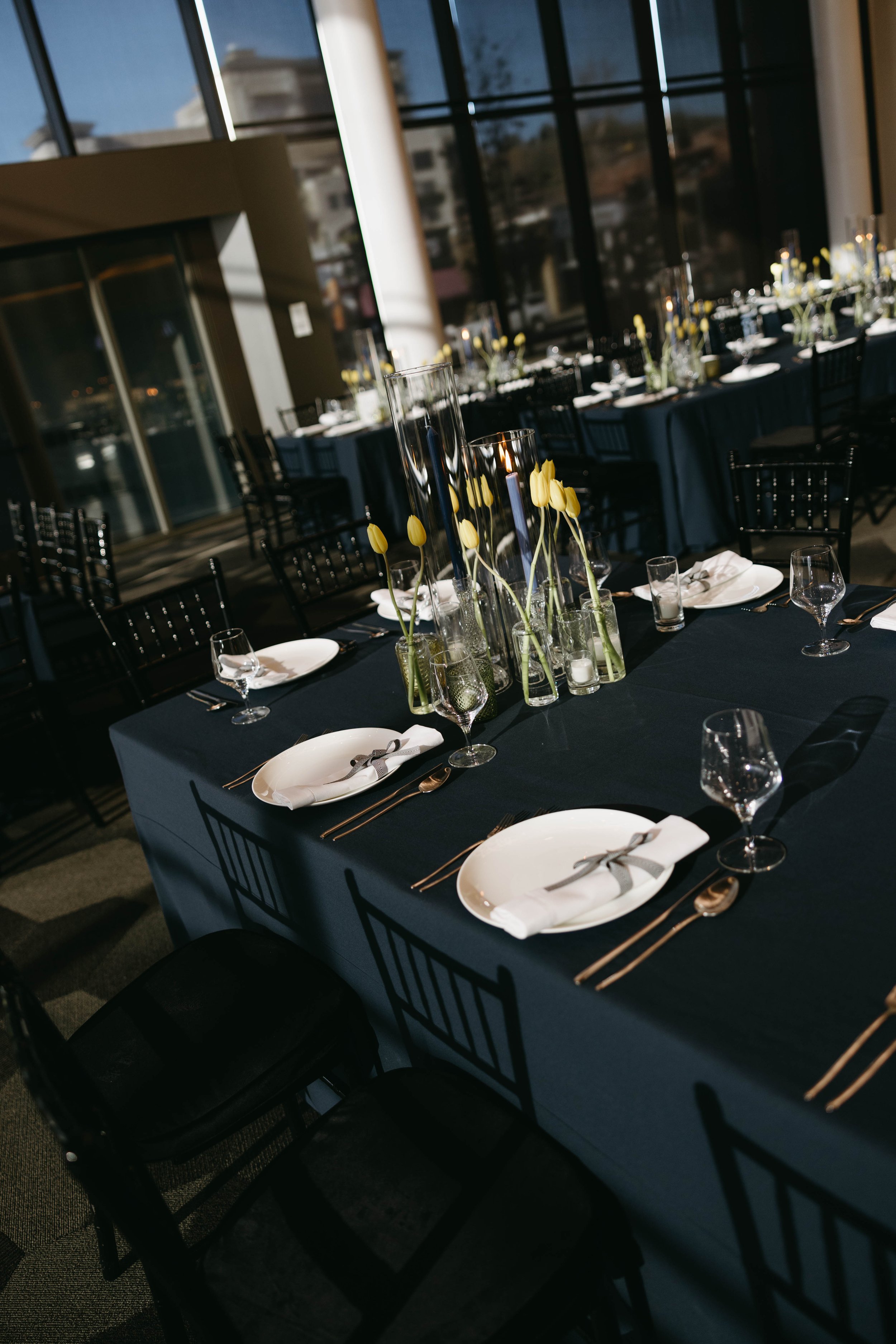 Elegant dining table setting with black chairs and dark tablecloths, decorated with yellow tulips in glass vases, white plates, utensils, wine glasses, and folded napkins. Large windows and blurred background visible.