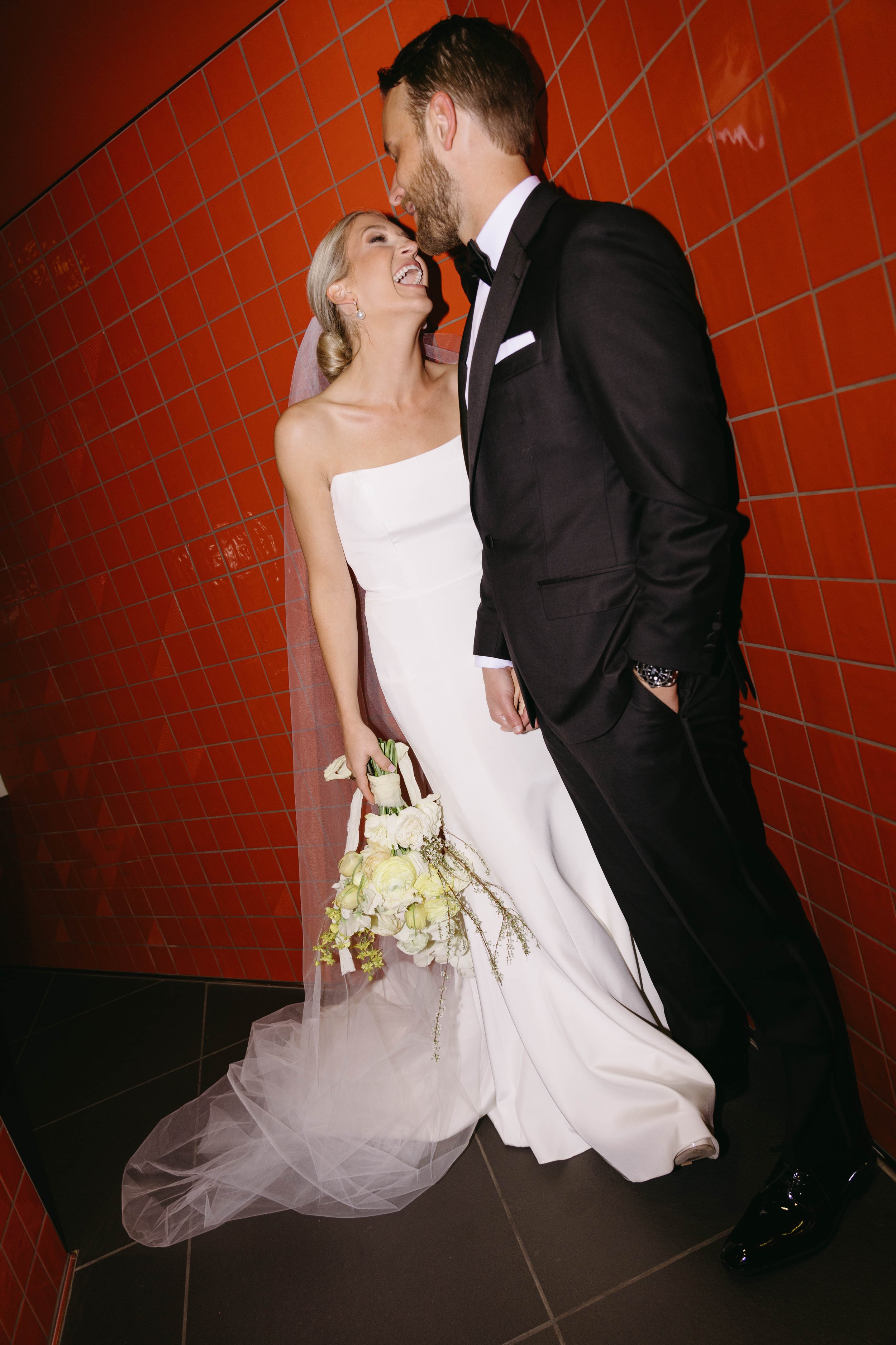 A bride in a white dress and a groom in a black suit are smiling at each other, standing in front of a red tiled wall. The bride is holding a bouquet of white flowers.