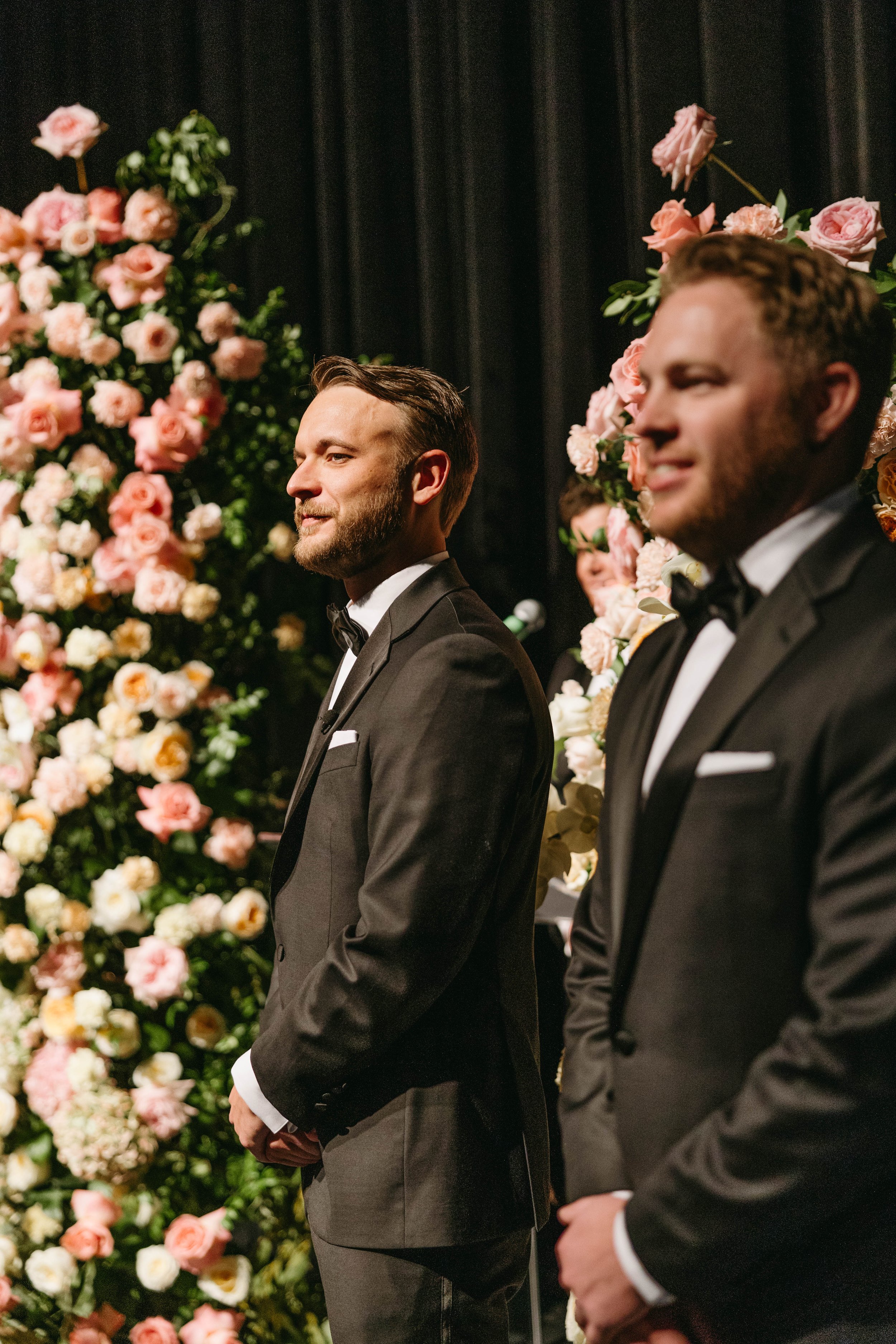 Two men in formal suits, standing in front of a floral arrangement with pink and white flowers, appearing to be in a wedding or formal event setting.