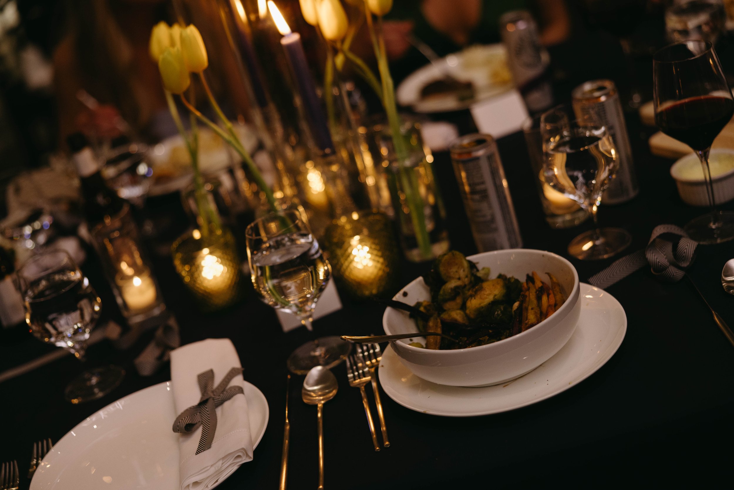 Elegant dining table set with bowls of food, wine glasses, yellow tulips, and candles.