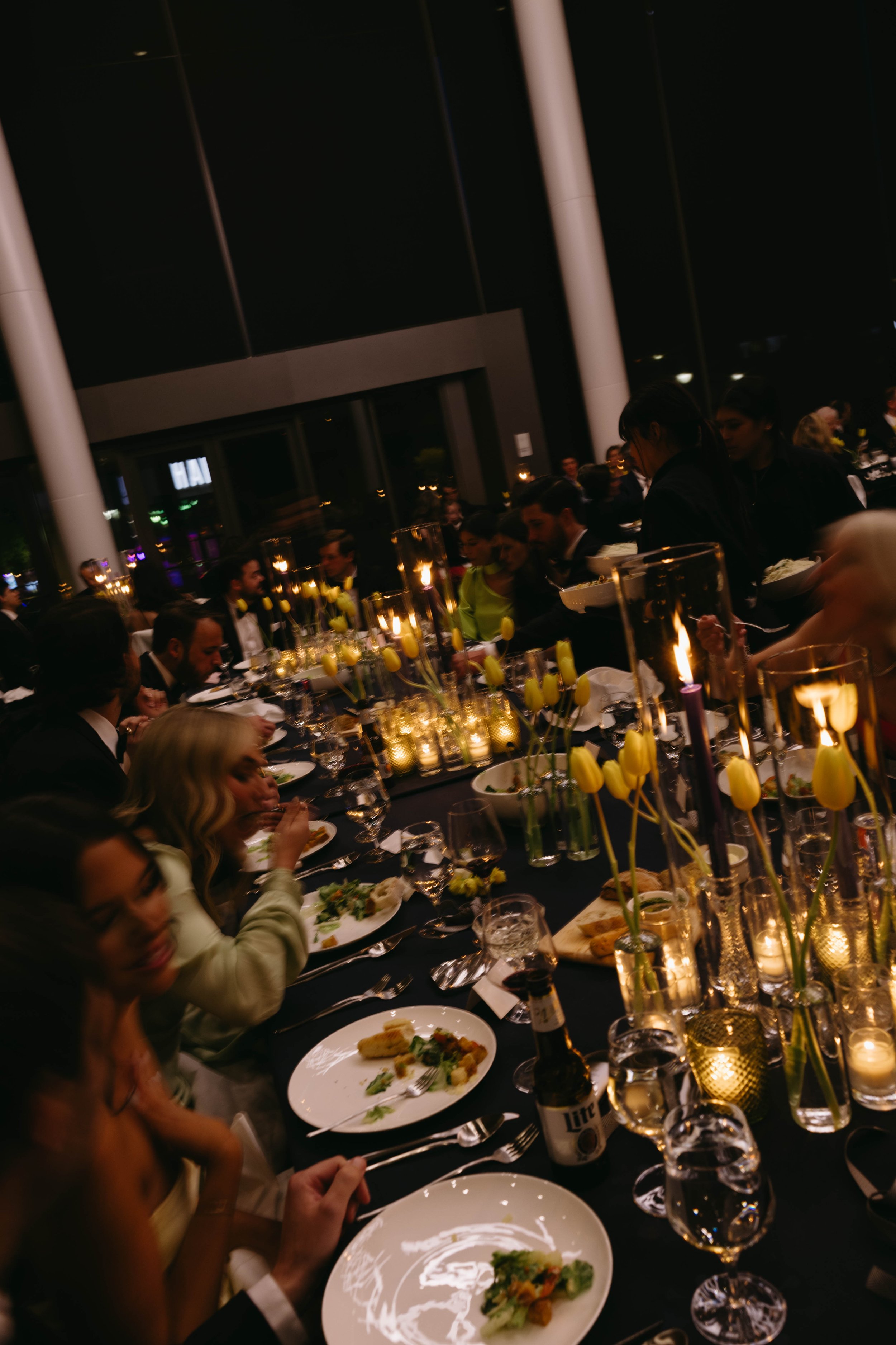 Elegant dinner event with guests seated at a long table, adorned with candles and yellow flowers, enjoying a meal. The setting appears formal with dim lighting and sophisticated decor.