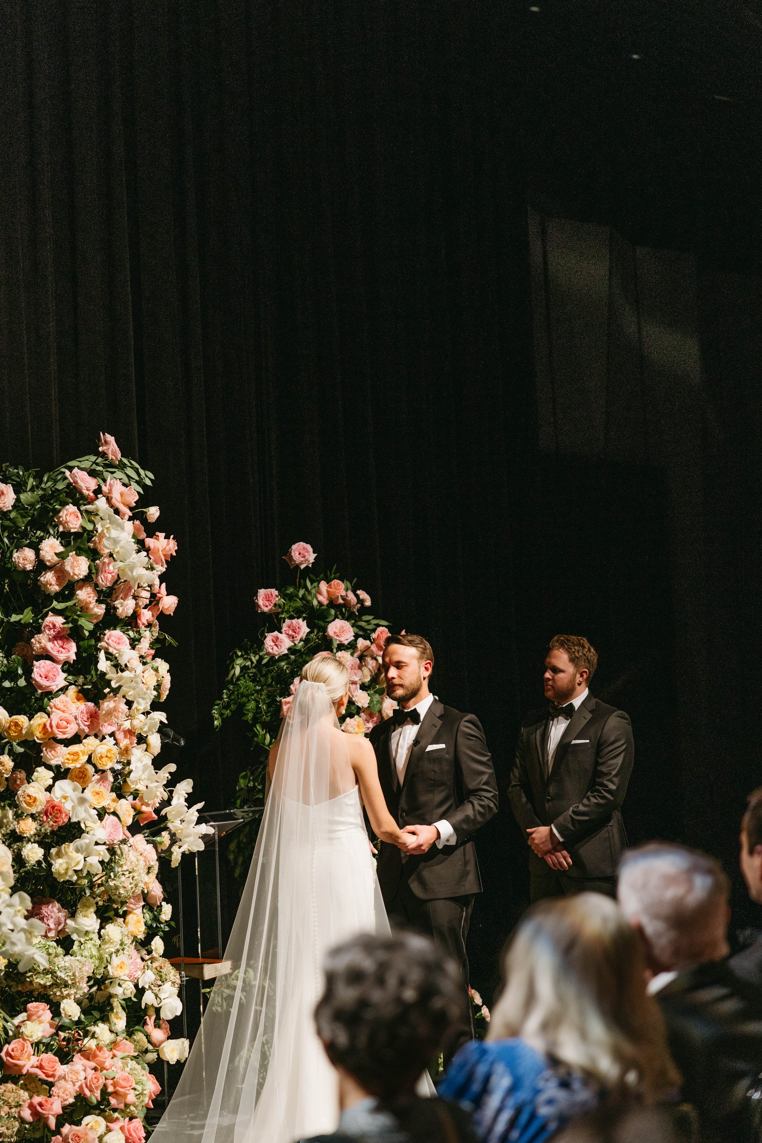 Bride and groom holding hands during wedding ceremony in front of floral arrangements, with best man standing nearby, attended by seated guests.