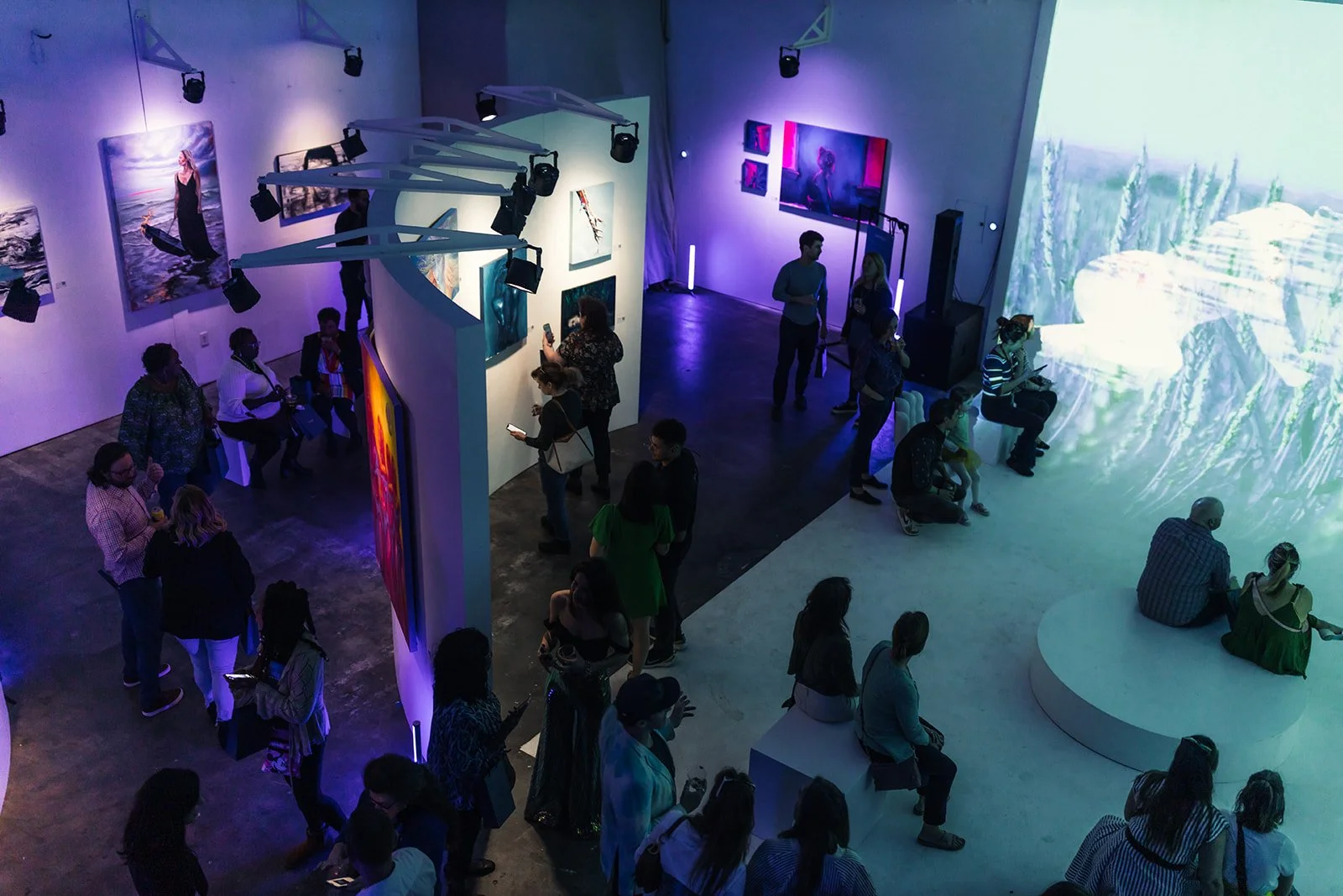 People attending an art gallery exhibition at Silver Hill Studio with various colorful paintings displayed on white walls, some viewing artwork and others sitting or standing near large digital screens projecting artistic visuals.