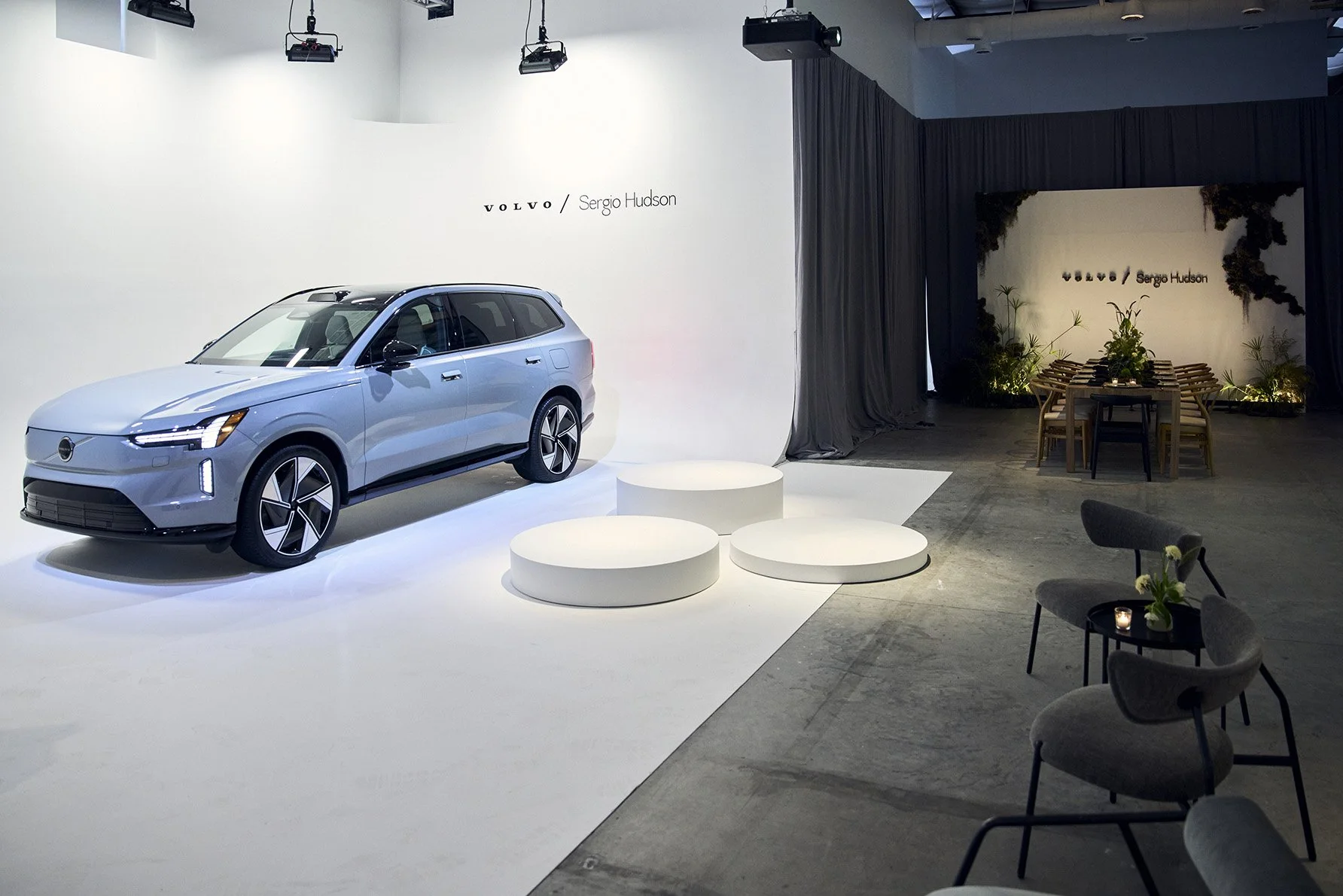White Volvo car displayed on a white platform at an indoor auto show with a backdrop that reads "Volvo Sergio Hudson." The setting includes minimalistic decor, with dark curtains, some chairs, and plants in the background.