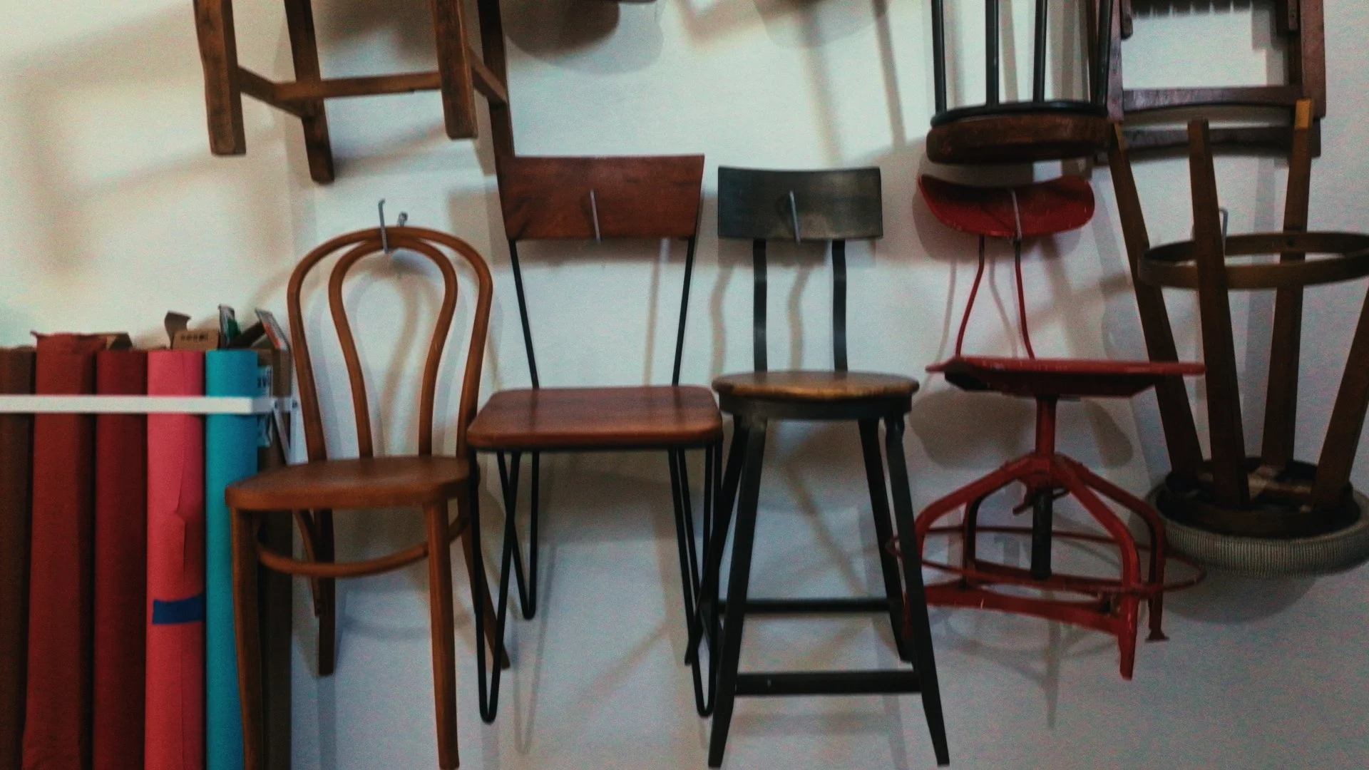 Collection of variously styled wooden and metal chairs arranged against a wall. Some chairs are stacked or leaning.