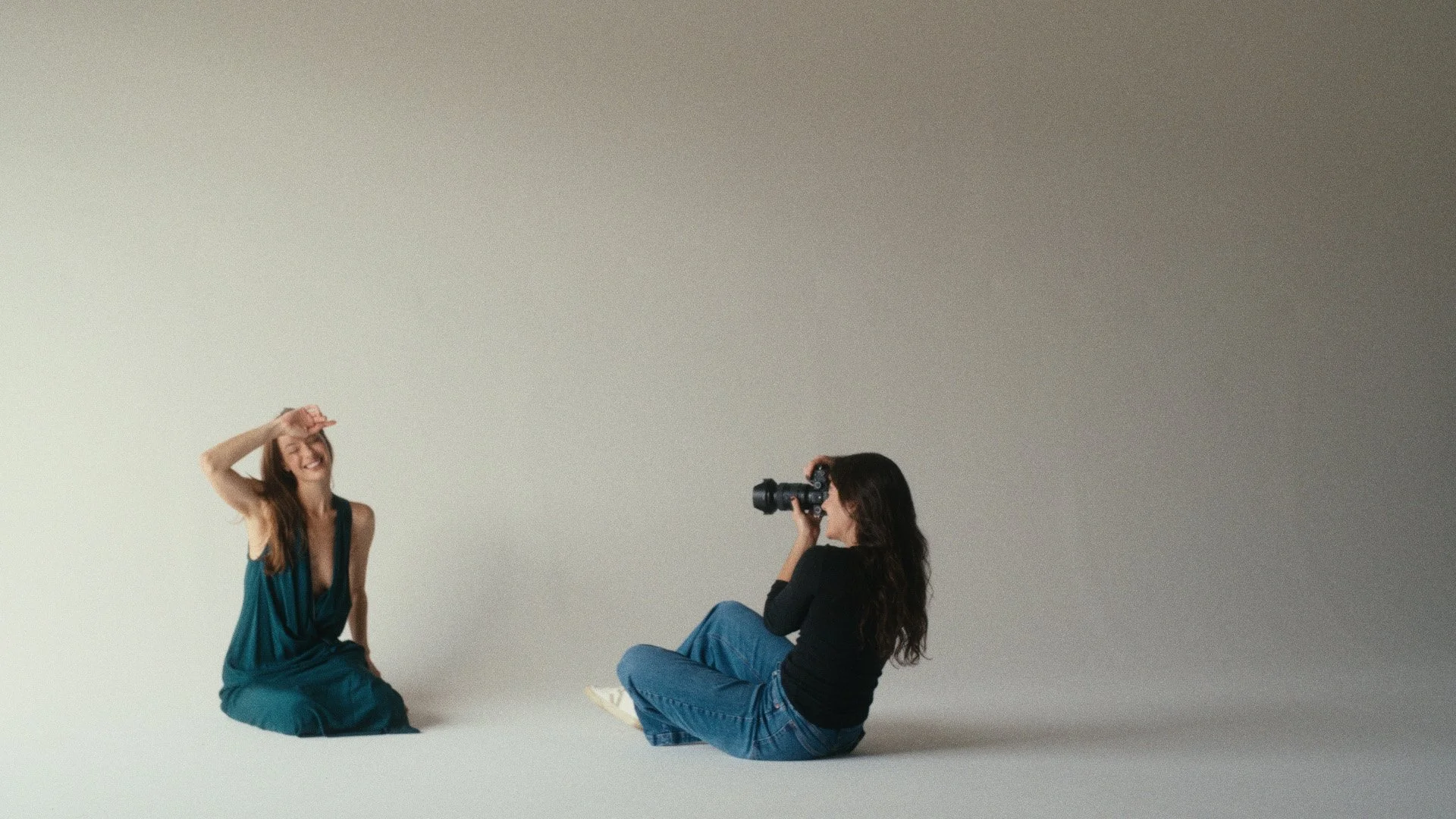 A female model sitting on the floor and smiling while a female photographer takes her photo with a camera against a plain white background showcasing Silver Hill Studio's two natural light areas.