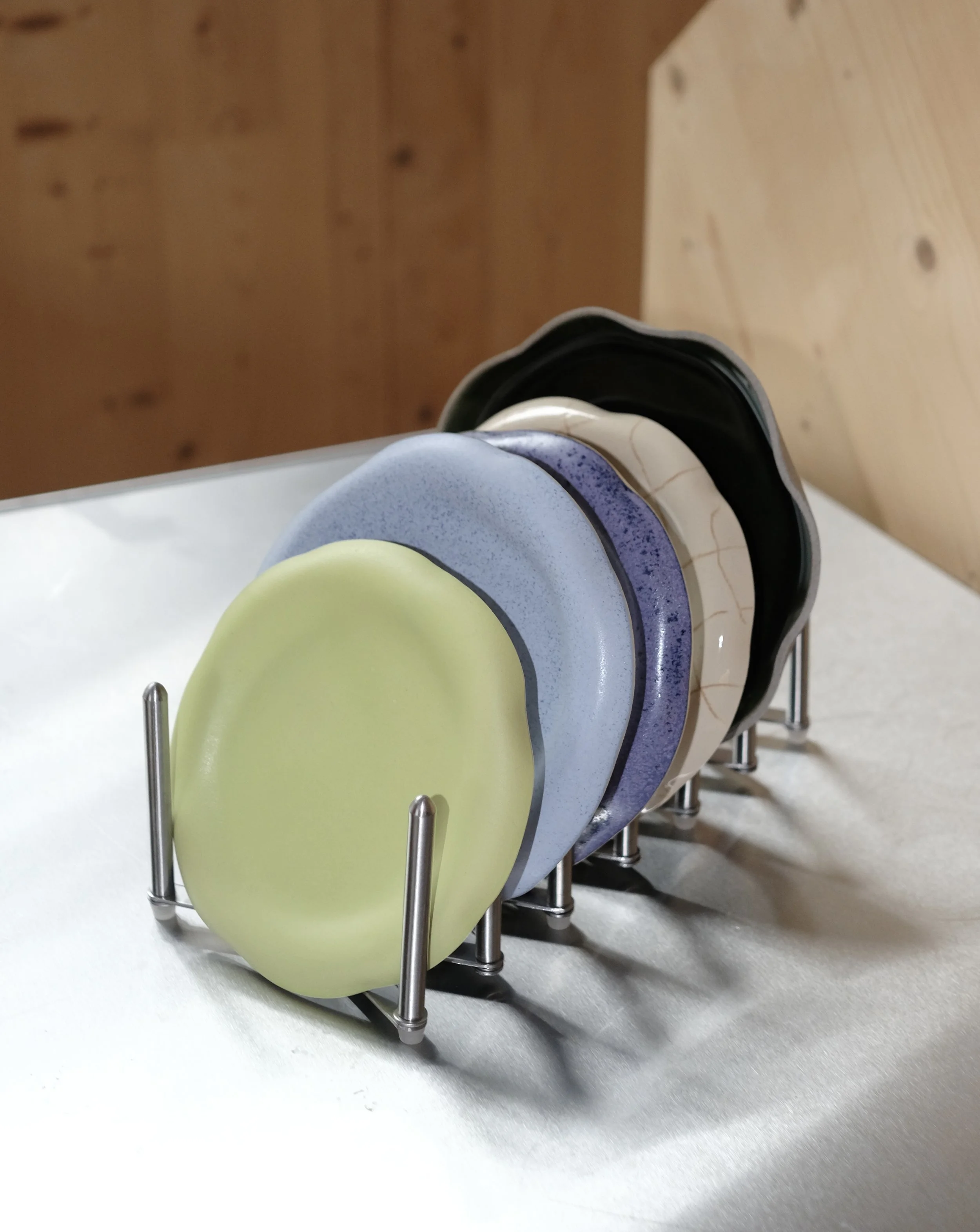 A stand holding seven colorful cstoneware plates in pastel shades, arranged in a row on a table with a wooden wall in the background.