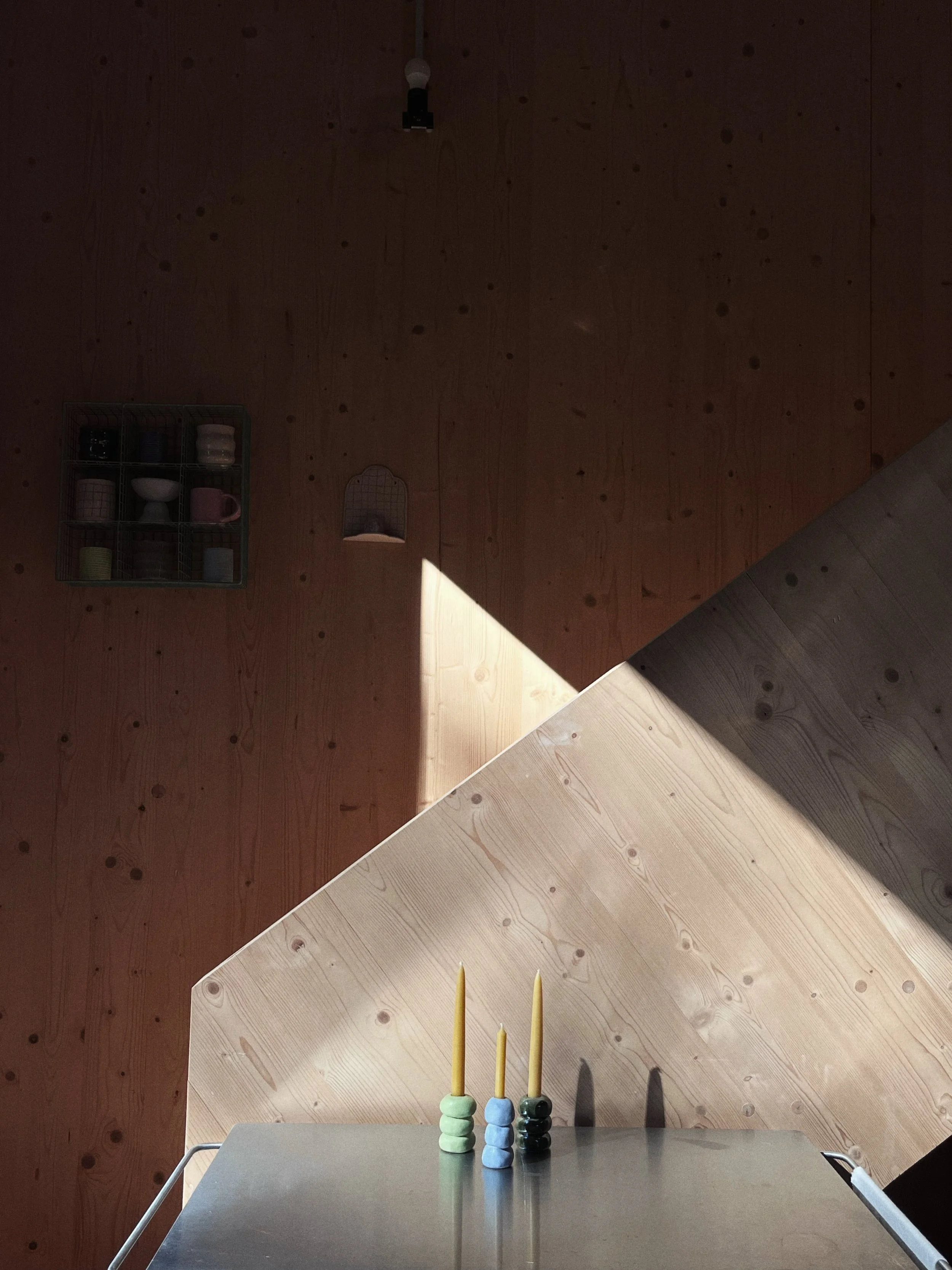 A minimalist interior with wooden walls and a metallic table holding three colorful candles with stacked holders.