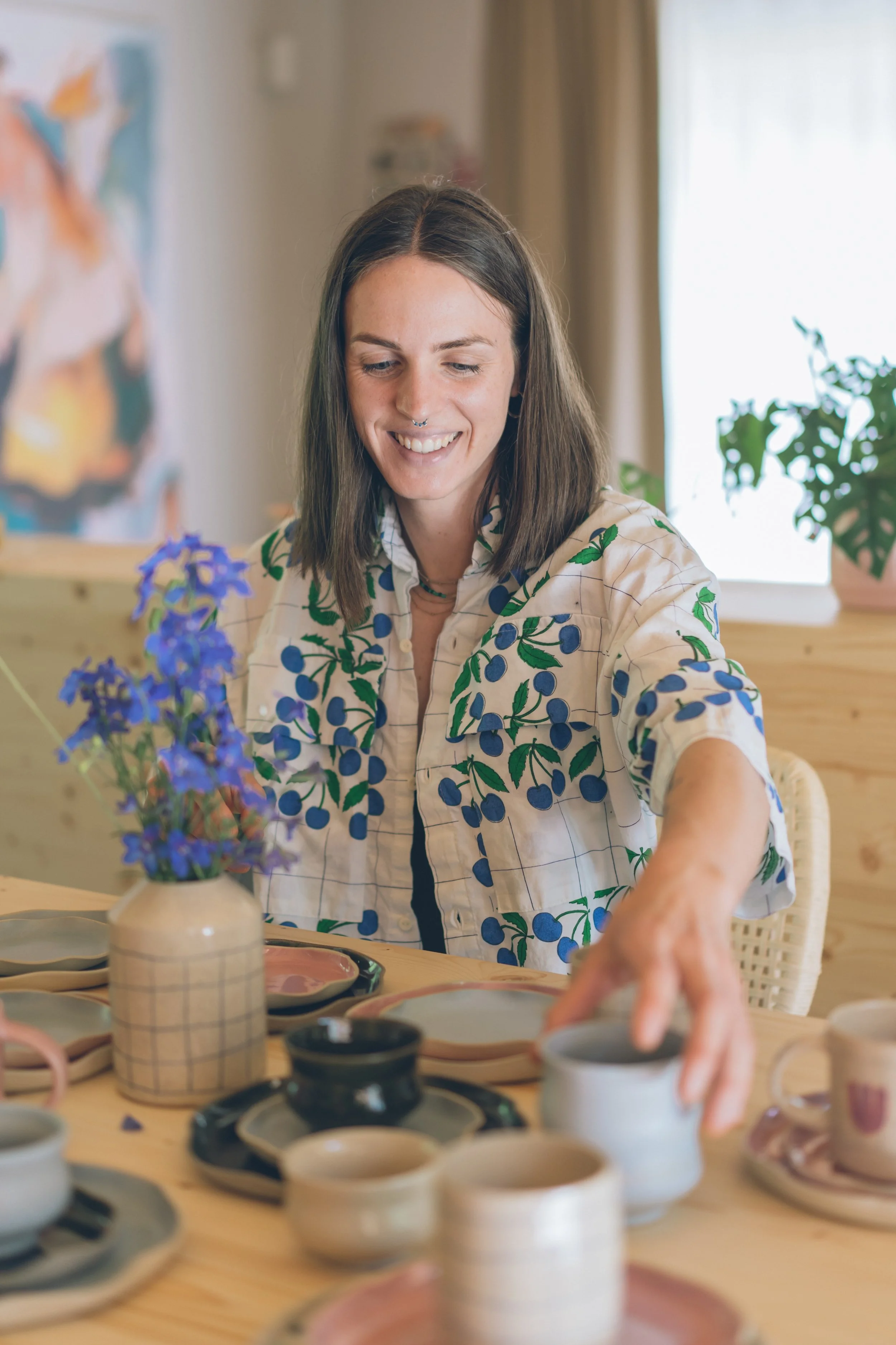 A woman with shoulder-length brown hair, wearing a white shirt with blue berries and green leaves, is arranging ceramic dishware on a wooden table, with purple flowers in a checkered vase nearby.