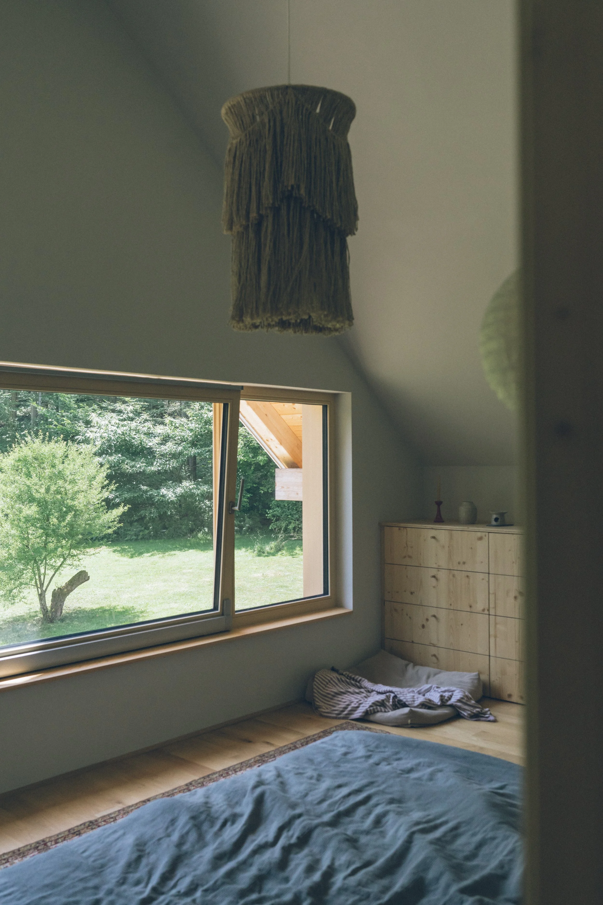 A cozy bedroom with a sloped ceiling, a large window showing a green backyard, a bed with light blue bedding, a wooden dresser in the corner, and a woven lampshade hanging from the ceiling.