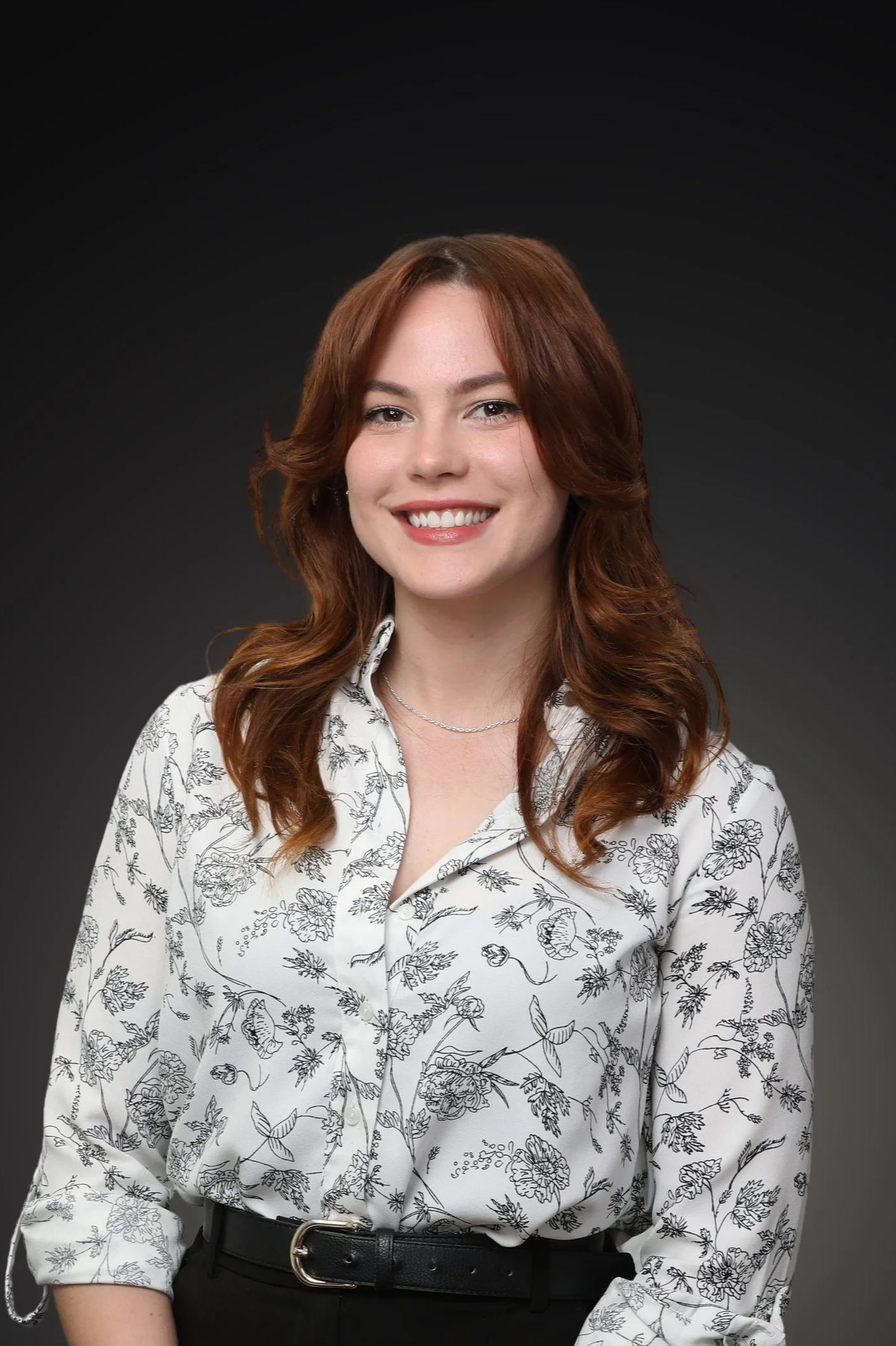 Close-up of a woman with long red hair, wearing a gray blazer, white shirt, pearl necklace, and gold hoop earrings, smiling against a dark background.