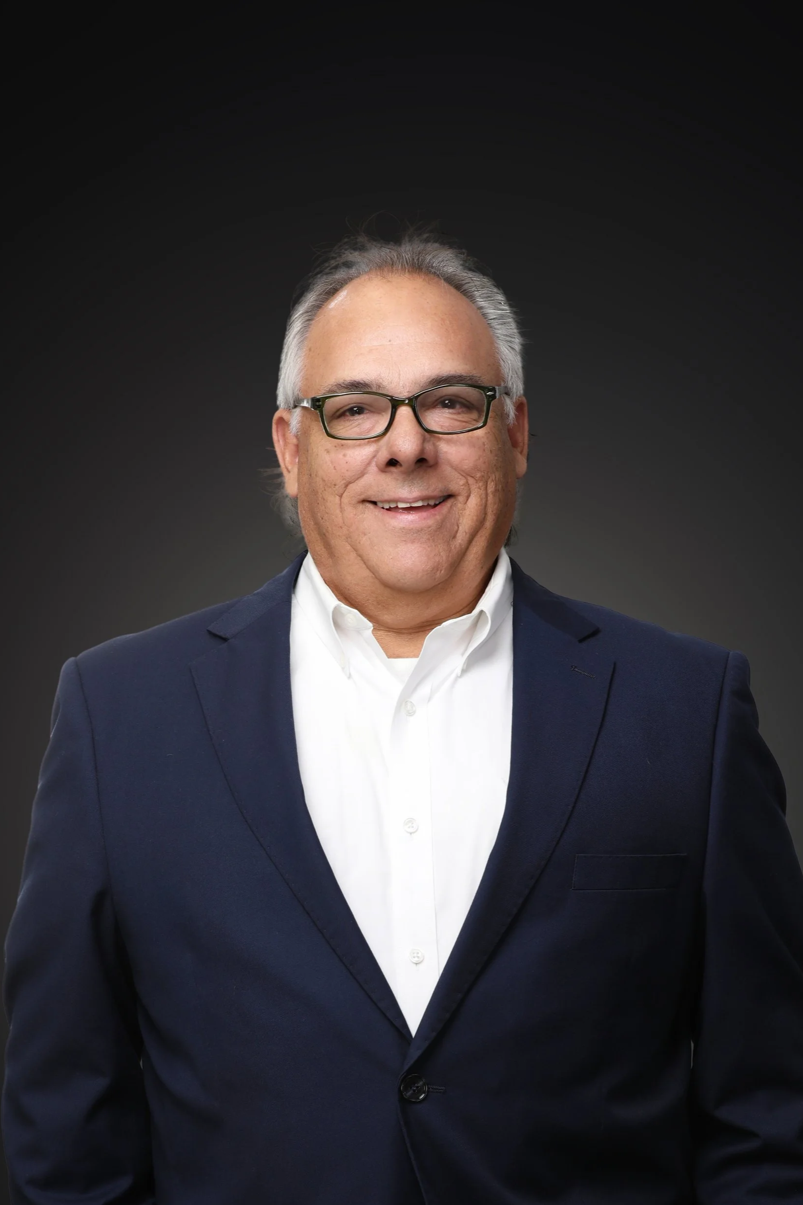 Close-up of a smiling man wearing glasses, a navy suit, and a white shirt, with a black background.