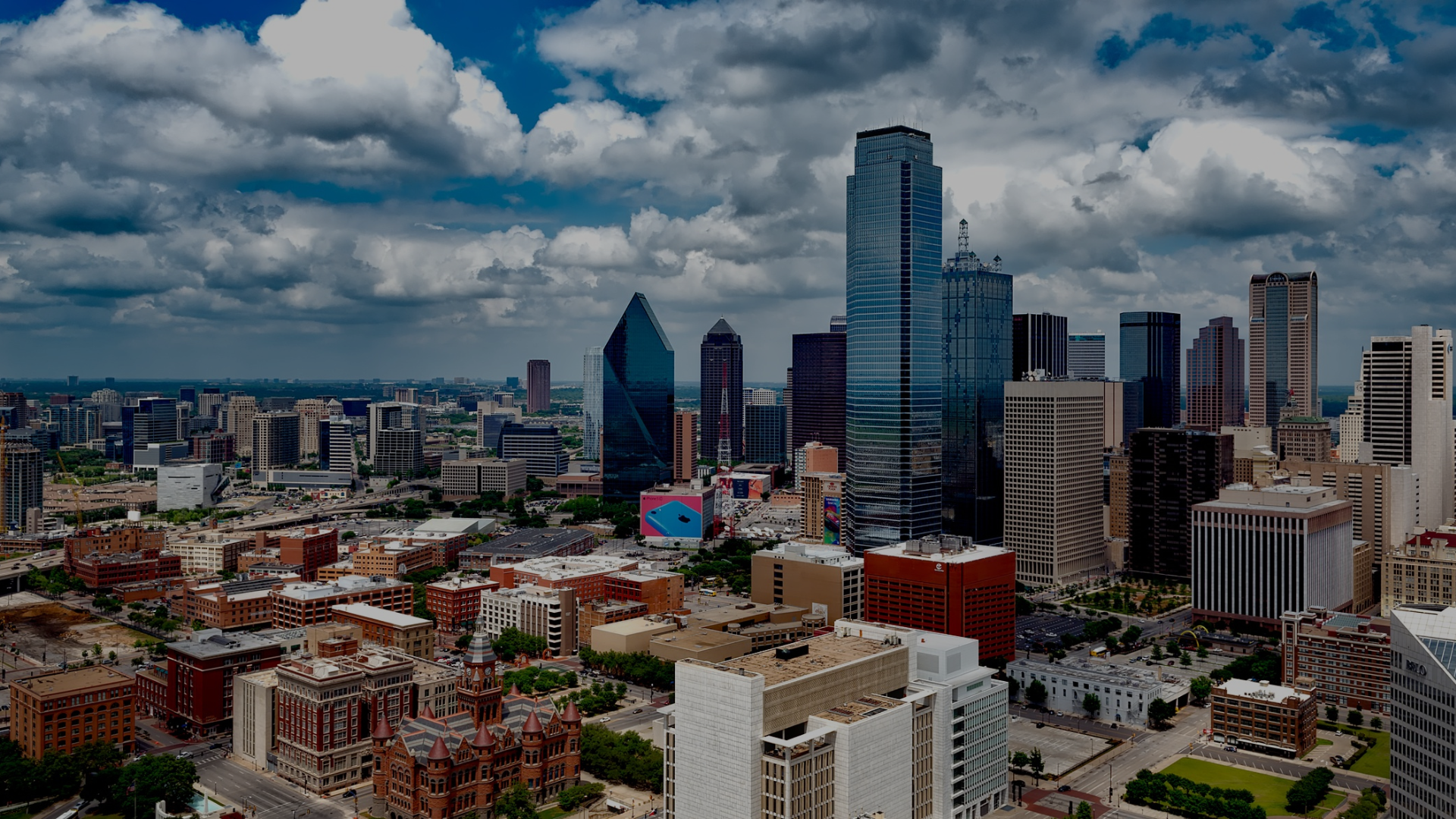 A city skyline with tall skyscrapers under a partly cloudy sky, including a prominent blue glass building and various other high-rise buildings and lower structures.