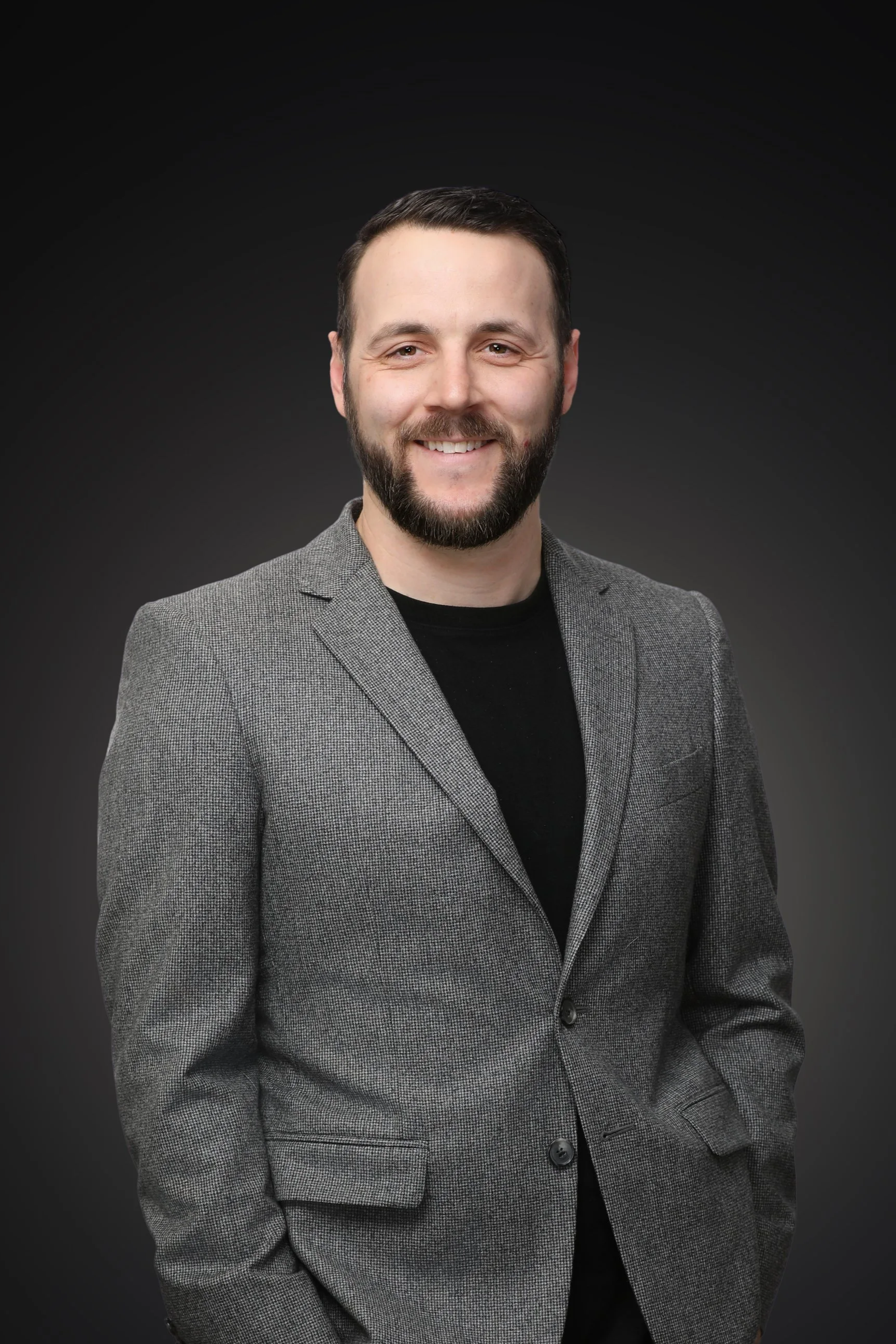 Close-up of a smiling man with dark hair, beard, and dressed in a black jacket against a dark background.