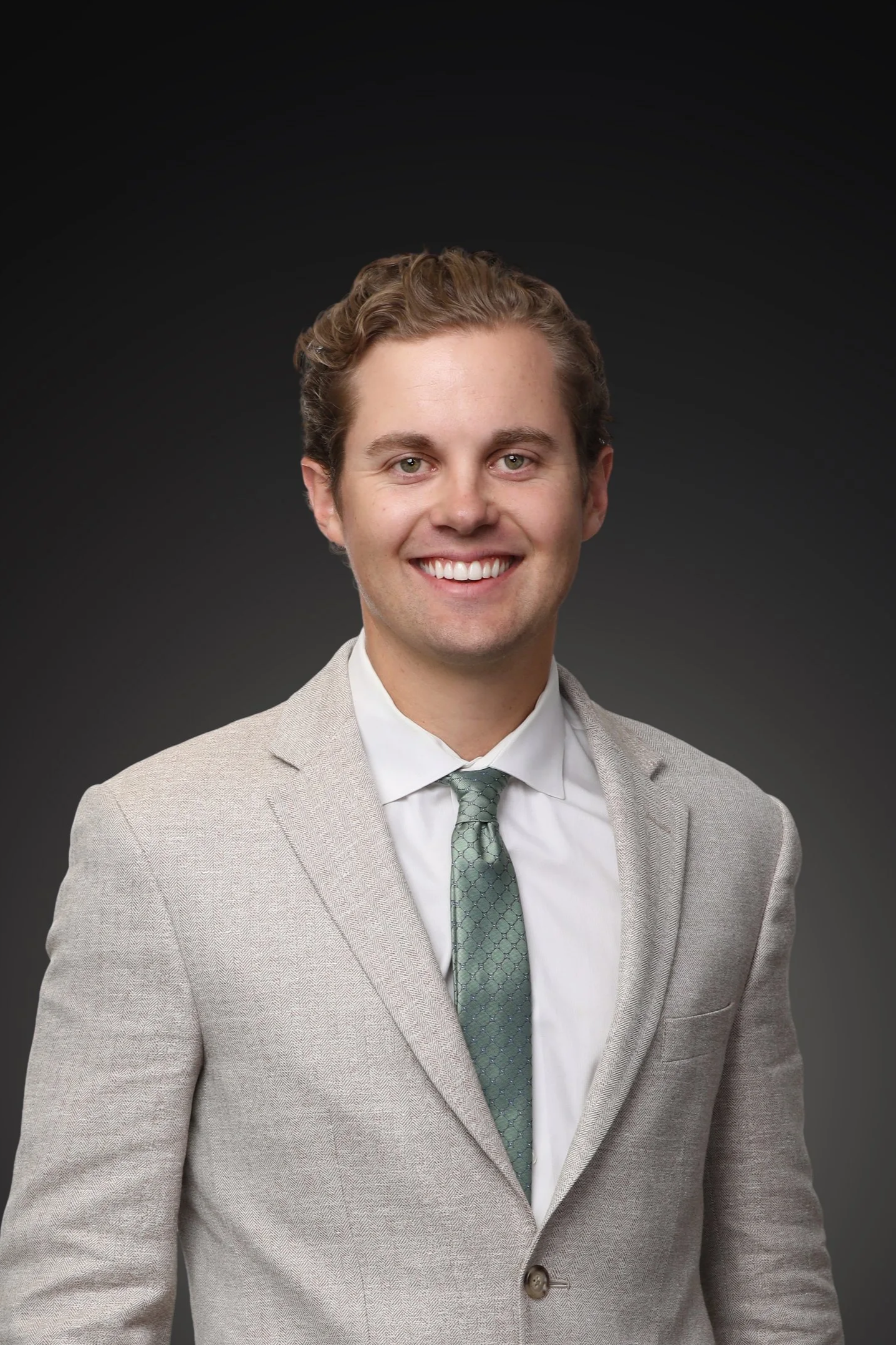 Close-up of a smiling young man's face, showing blue eyes, a well-groomed beard, and curly light brown hair, dressed in a beige blazer, white shirt with red checks, and a black tie, against a gray background.