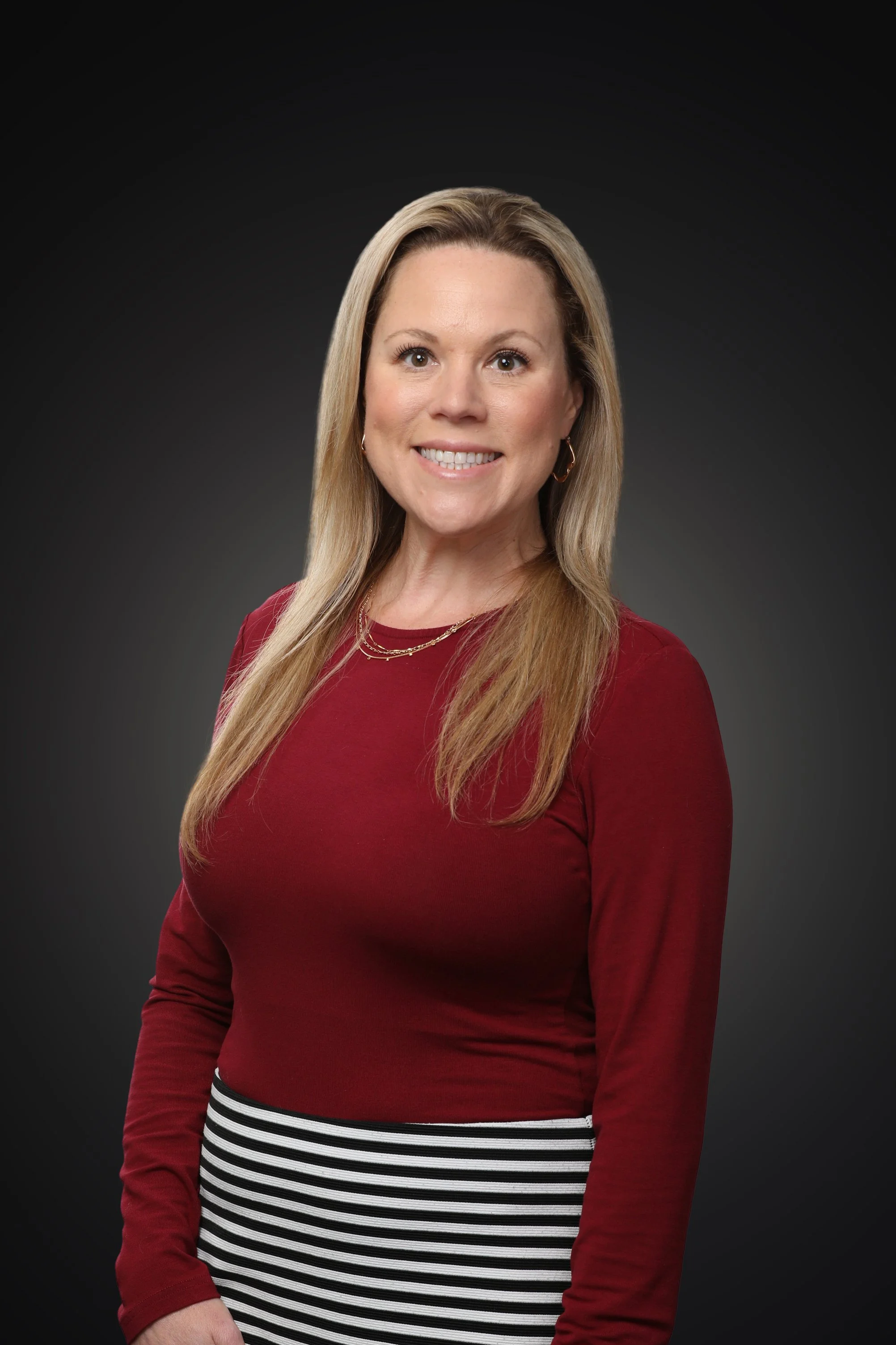 Close-up portrait of a smiling woman with long blonde hair, wearing a black top and a dark red blazer against a dark background.