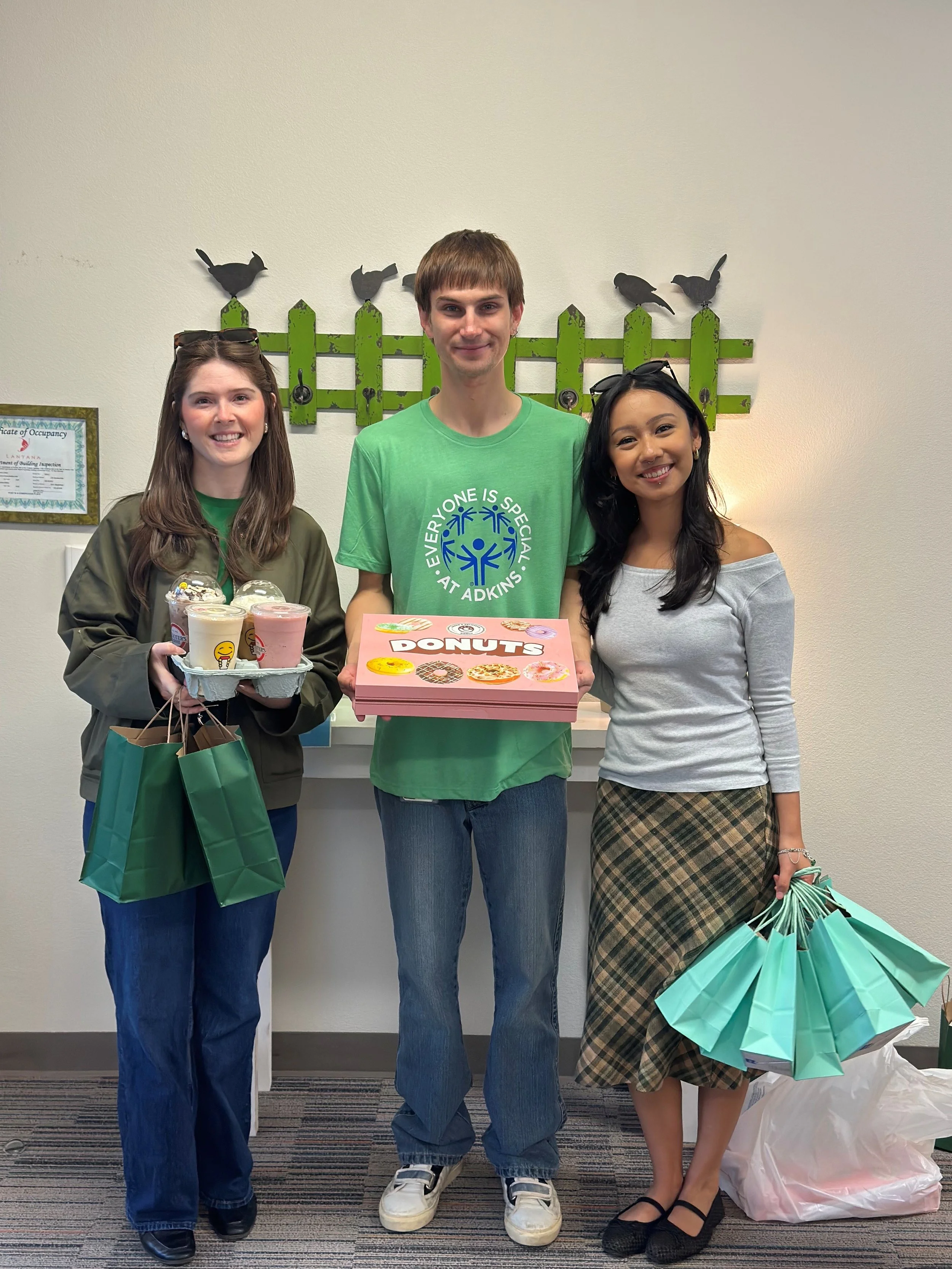 Three people standing together, holding donuts and shopping bags, in a room with a green fence and bird decorations on the wall.